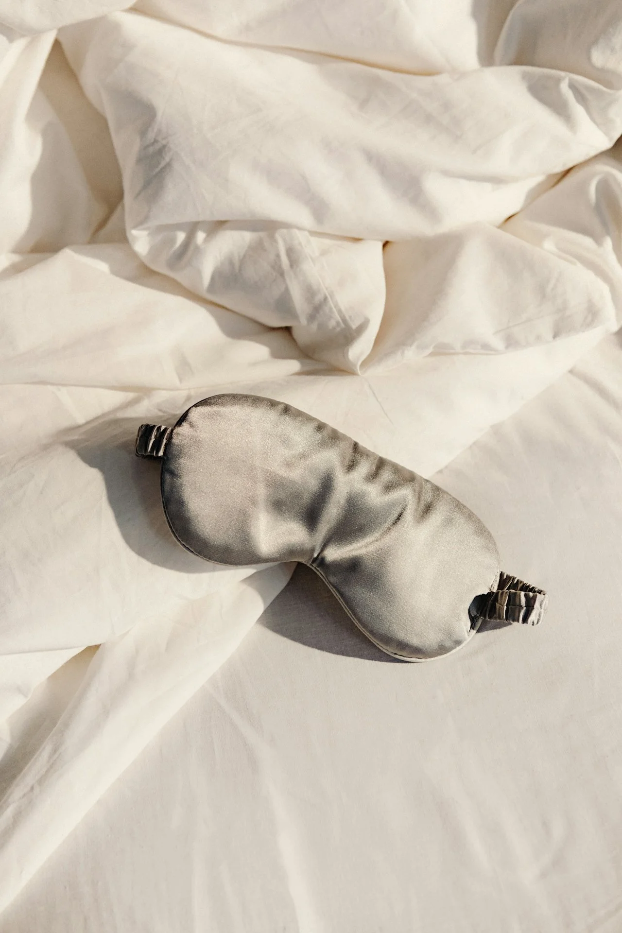 A satin eye mask with an elastic band, lying on white bedsheets, with crumpled white fabric in the background.