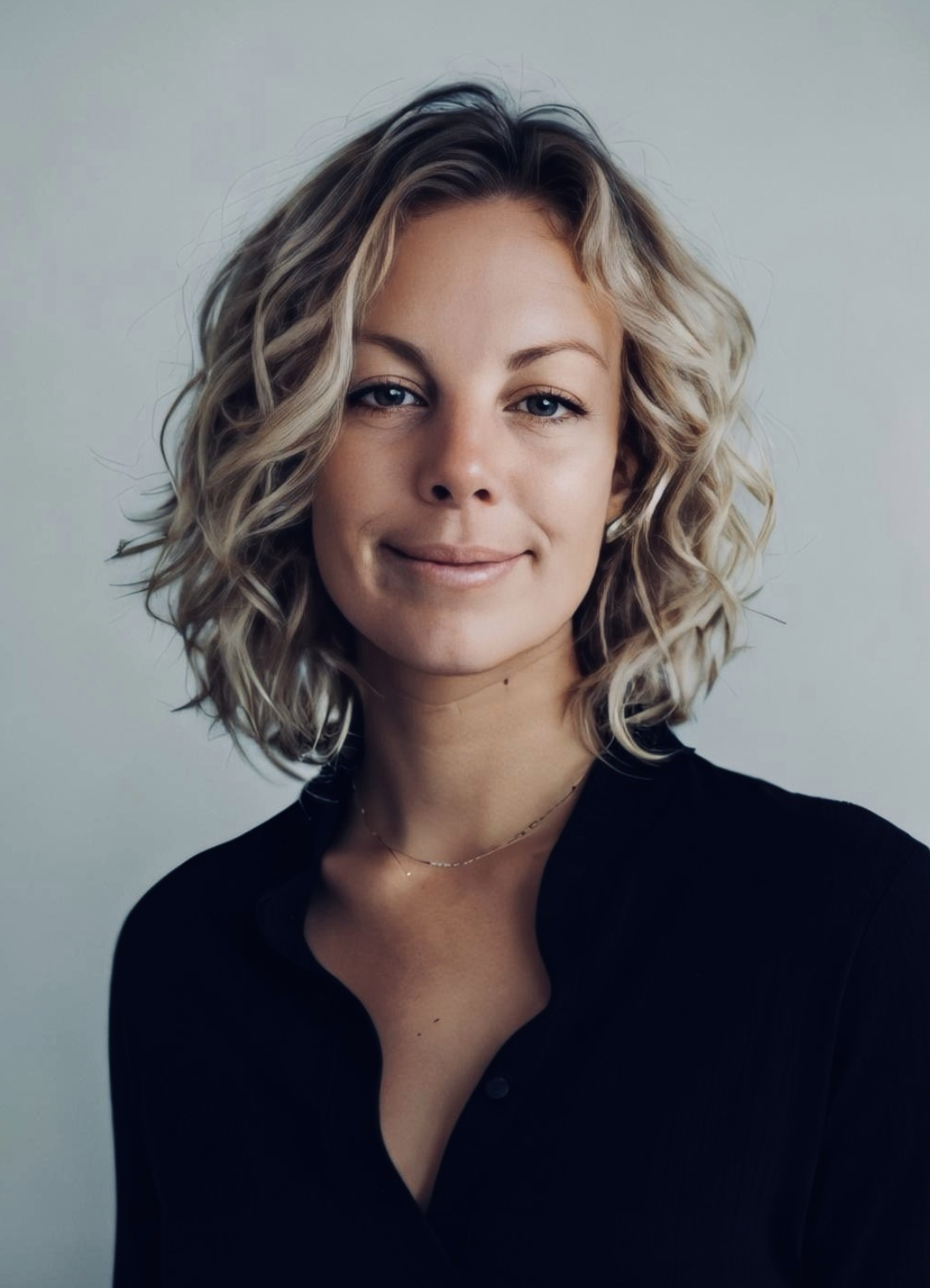 Close-up portrait of a woman with curly blonde hair, wearing a black top and a delicate necklace, smiling softly against a plain background.