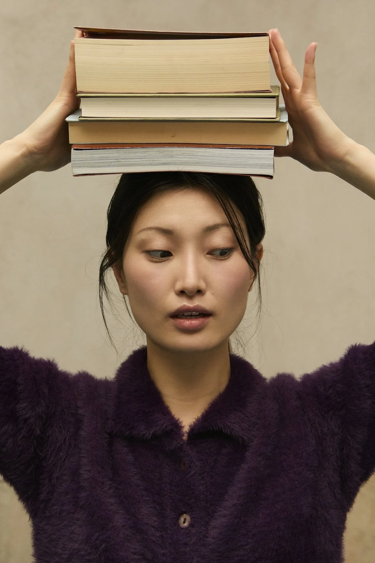 A woman with dark hair wearing a purple fuzzy coat balancing four books on her head, holding them with hands on both sides, against a plain background.