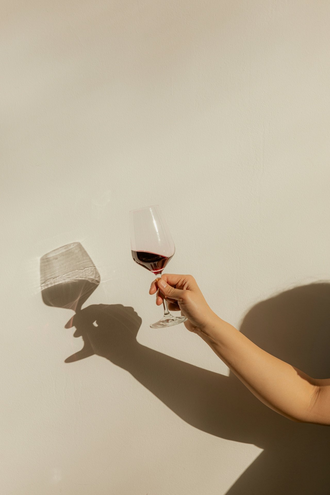 A person holding a glass of red wine against a plain white wall, casting a shadow.