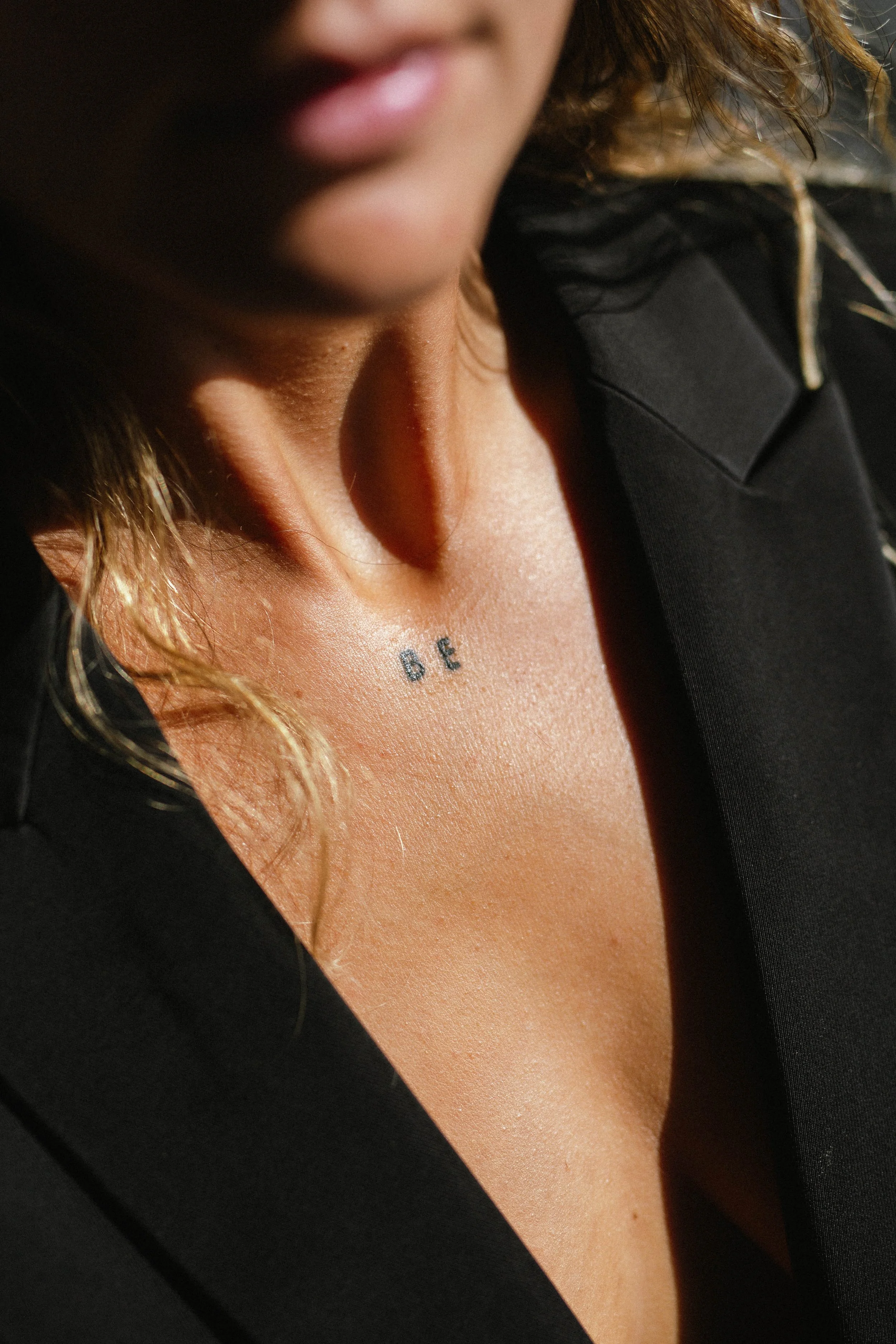 Close-up of a woman's neck with a tattoo that reads 'BE', wearing a black shirt, and with curly hair visible.