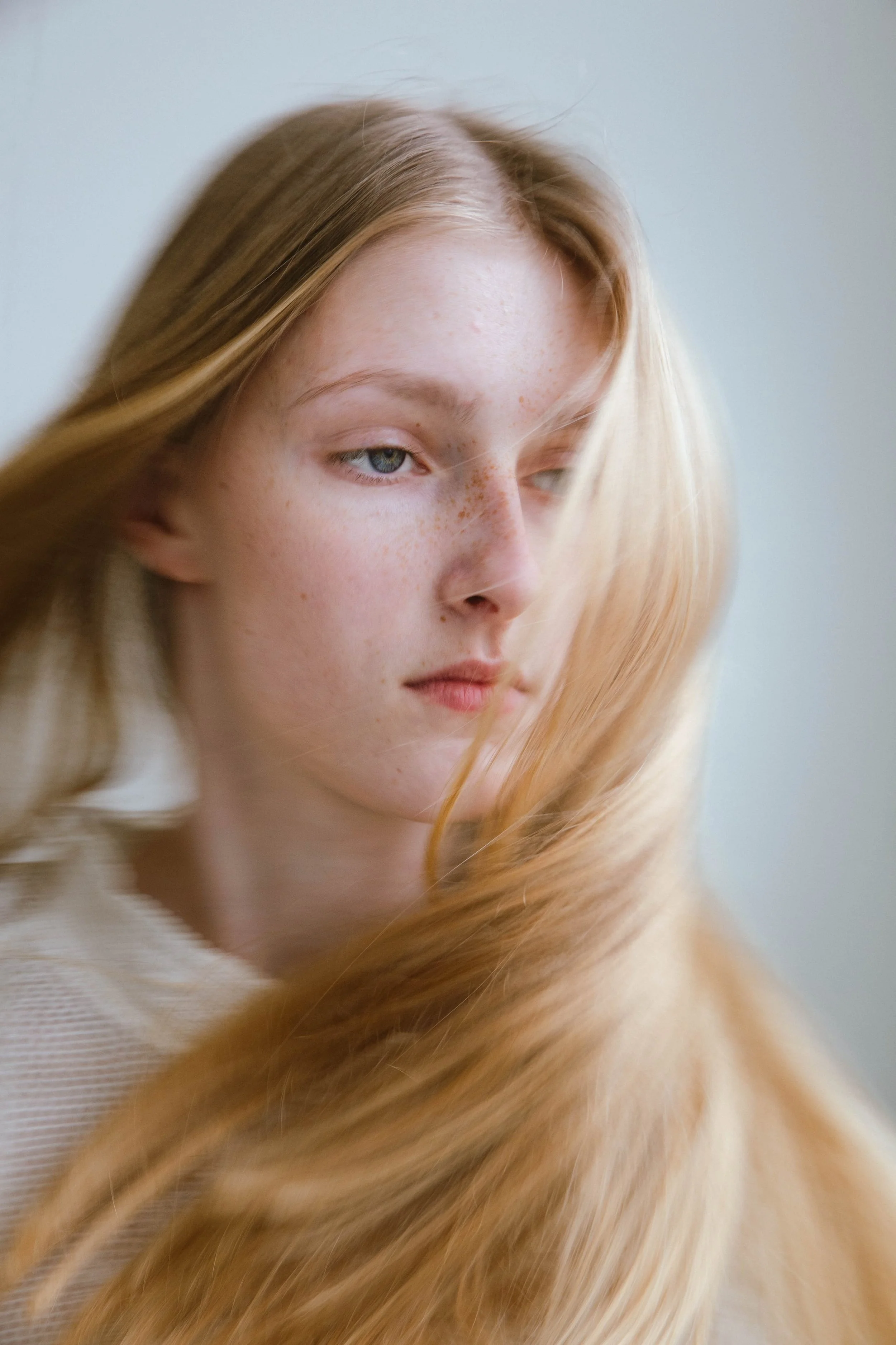 Close-up of a young woman with long, flowing red hair and light skin with freckles, looking slightly to the side against a neutral background.