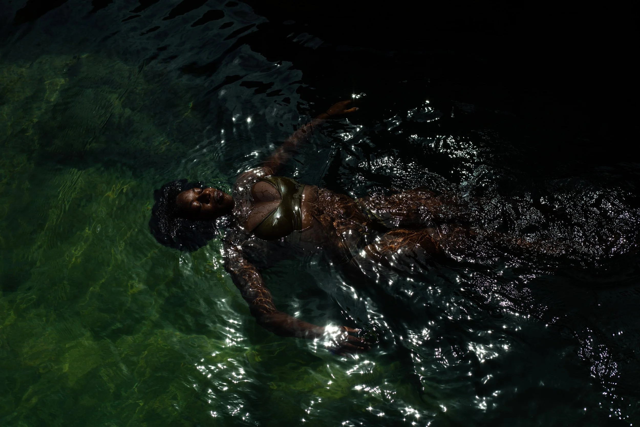 A woman floating in dark water with a serene expression on her face, wearing a green swimsuit, with sunlight reflecting off the water surface.