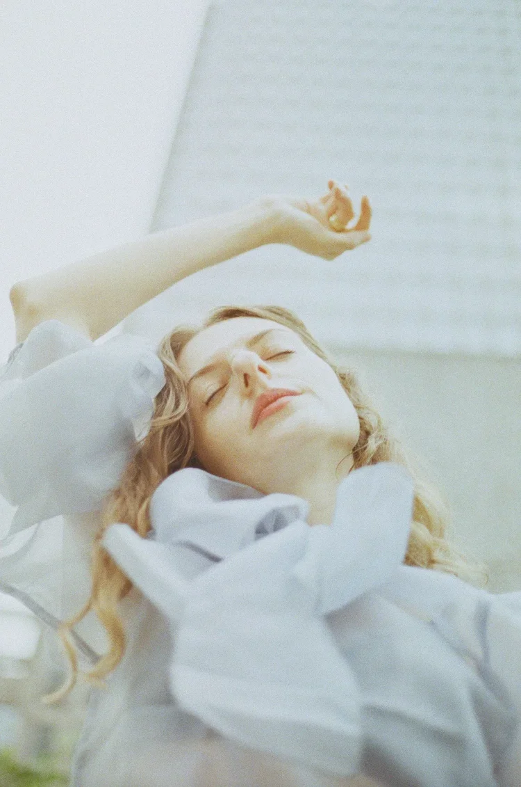 A woman with curly blonde hair lying peacefully with her eyes closed, wearing a white shirt, beside a body of water.