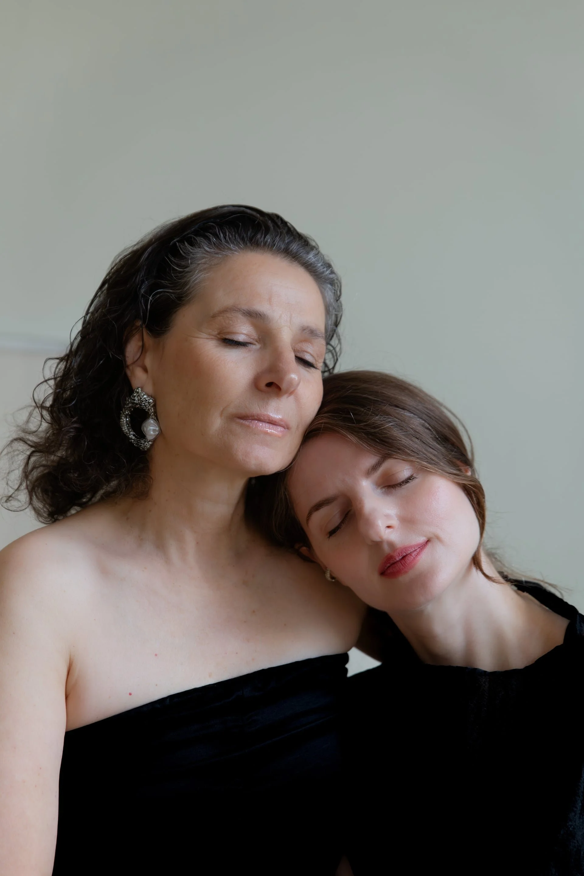 A woman with dark, curly hair and earrings gently embracing a young girl with straight, light brown hair and closed eyes, both wearing black off-the-shoulder tops and appearing peaceful.