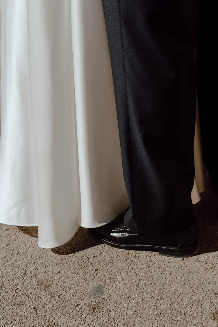 Close-up of a person's bottom half wearing black dress shoes, black pants, next to a white dress or robe on a tan carpeted floor.