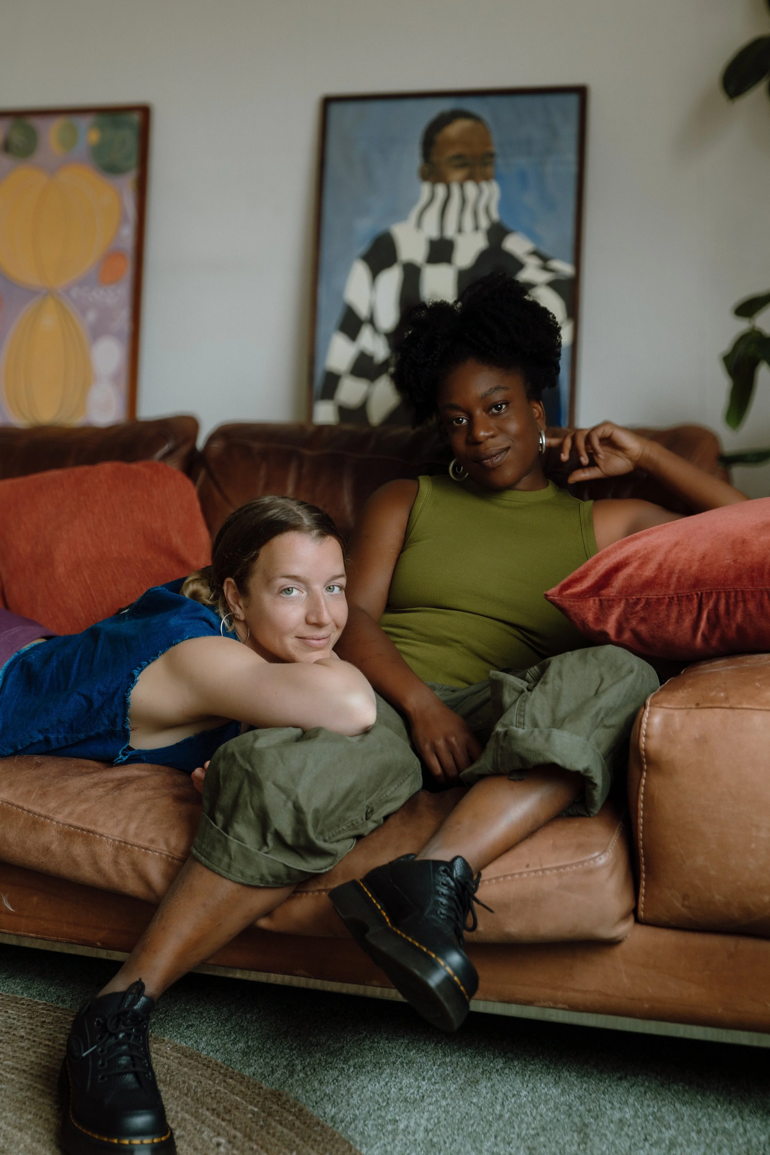 Two women sitting on a brown leather sofa in a living room. One woman with curly hair in an olive green tank top, and the other with straight hair in a blue top. Behind them are colorful abstract paintings and a houseplant.