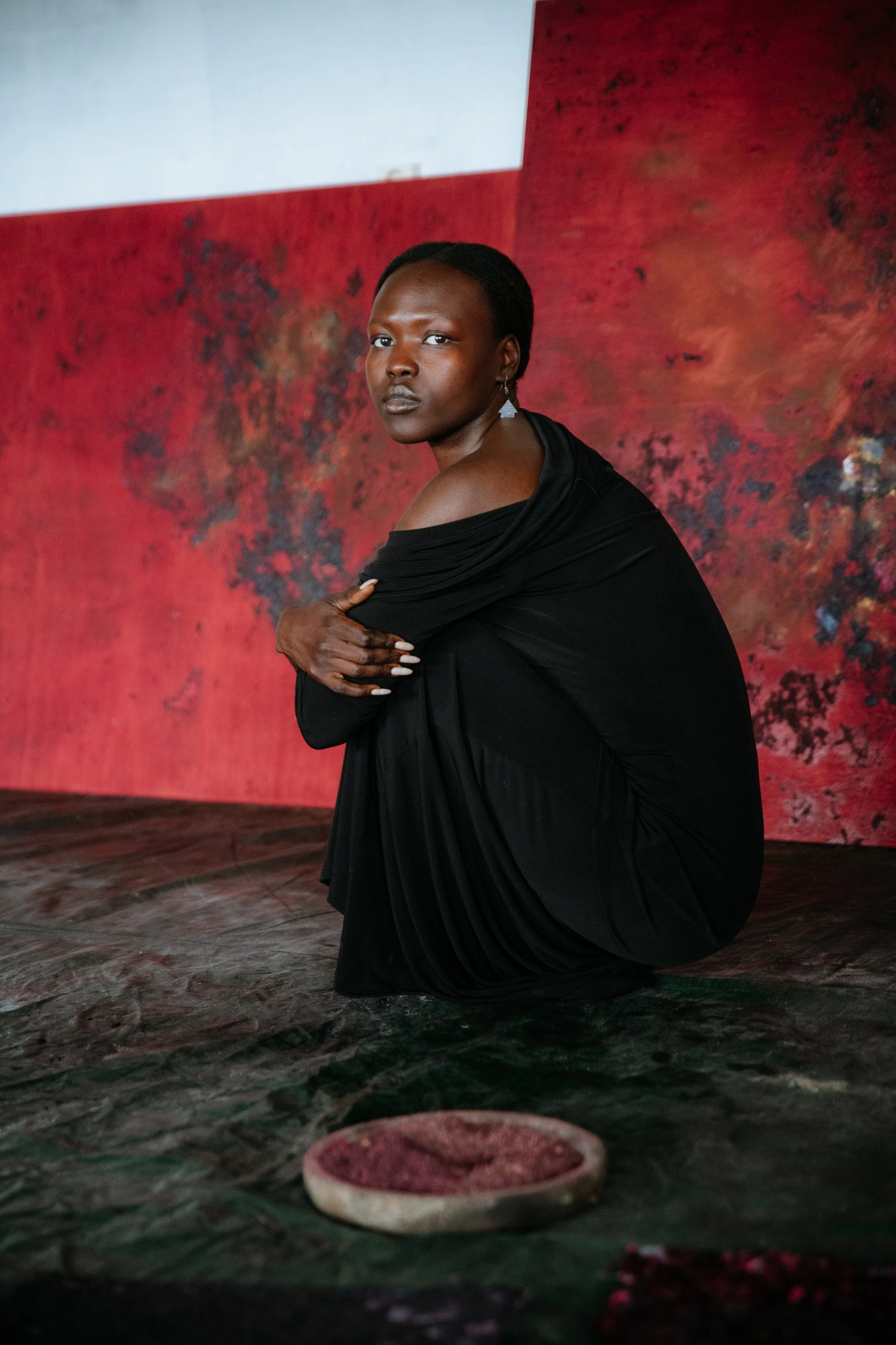 A woman with dark skin and short hair, dressed in black, kneeling on the ground in front of a textured red background, looking directly at the camera.