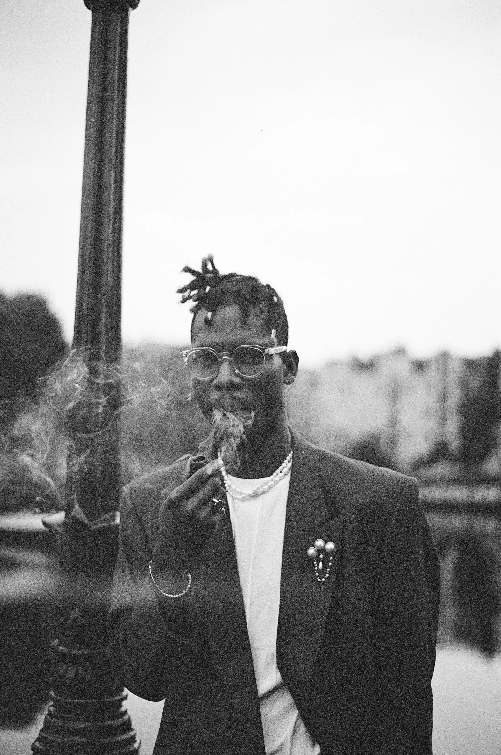 A person with dreadlocked hair, wearing sunglasses, a pearl necklace, and a suit jacket, is smoking a cigarette outdoors. There is a telephone pole in the background and some blurred foliage.