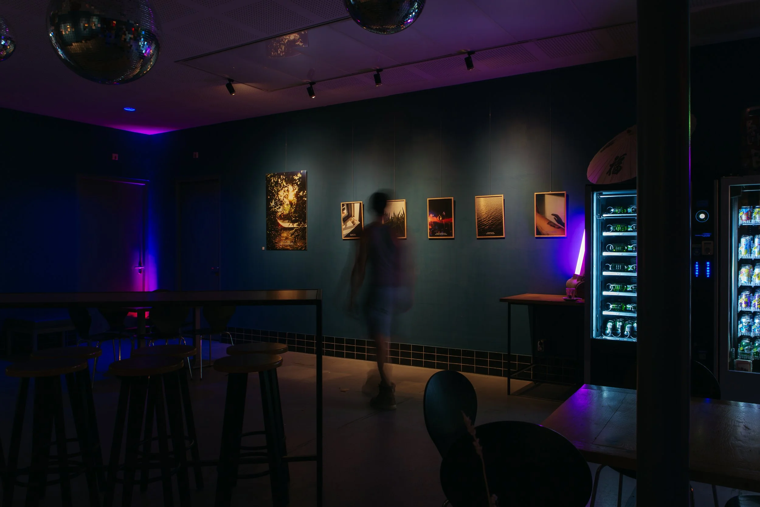 A dimly lit indoor space with dark walls, showing a person walking past a series of framed photographs on the wall, and a vending machine with illuminated shelves on the right side of the image.