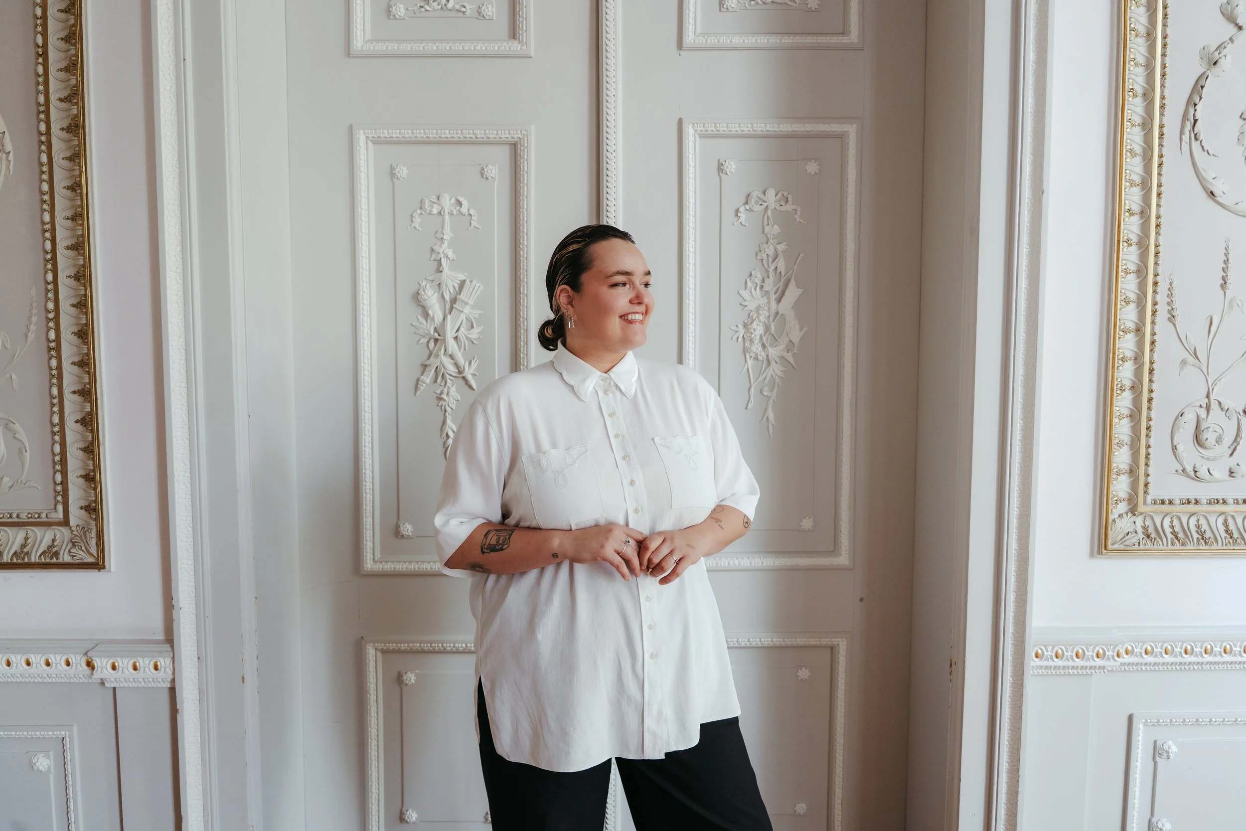 A woman with dark hair styled in a low bun wearing a white button-up shirt with rolled-up sleeves, standing in front of an ornate white wall with decorative panels and gold accents, smiling and clasping her hands in front of her.