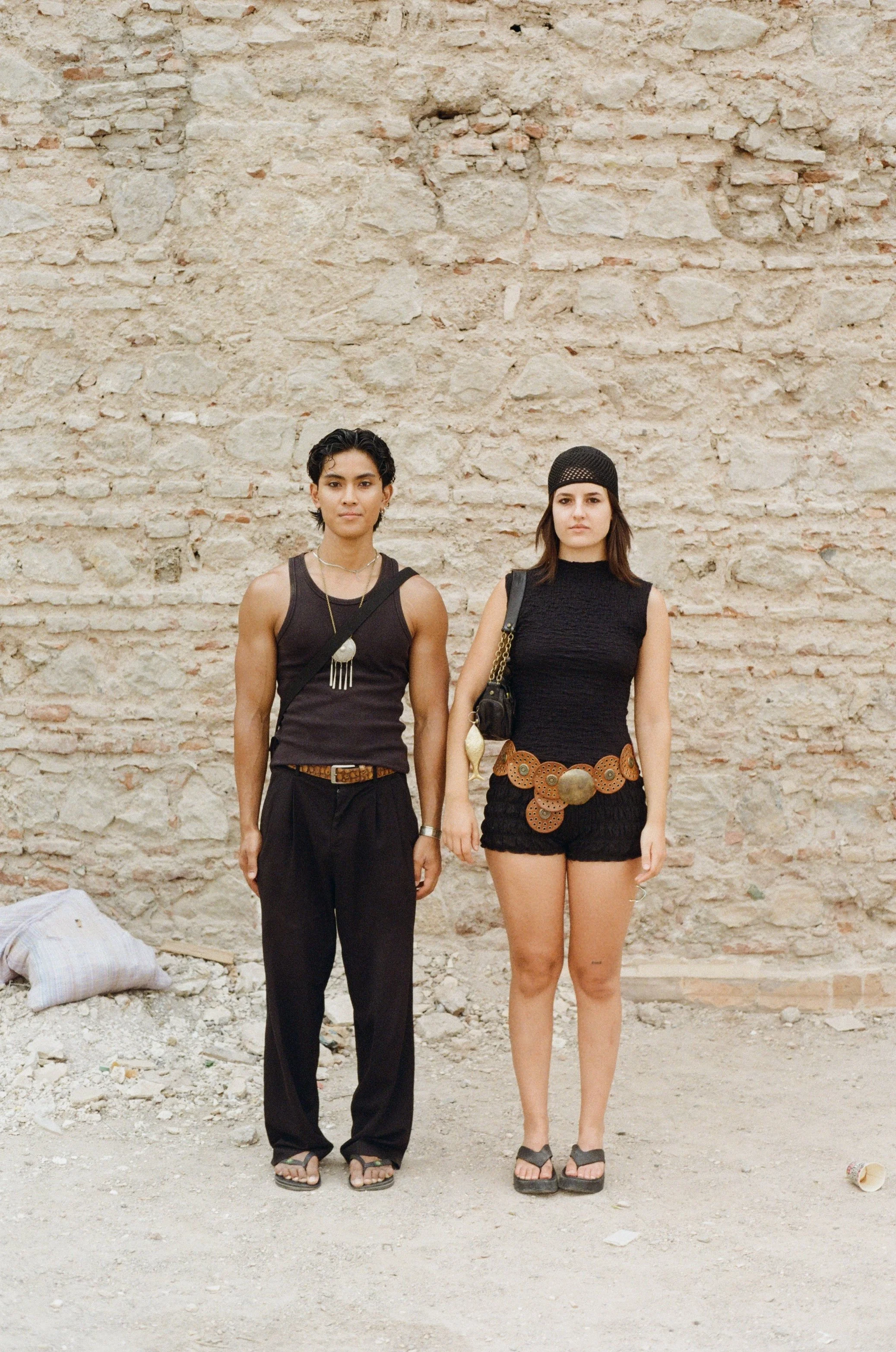 Two women standing in front of a brick wall, one in a sleeveless black top and black pants with dark hair, and the other in a sleeveless black dress with a hat and brown belt, both wearing sandals.