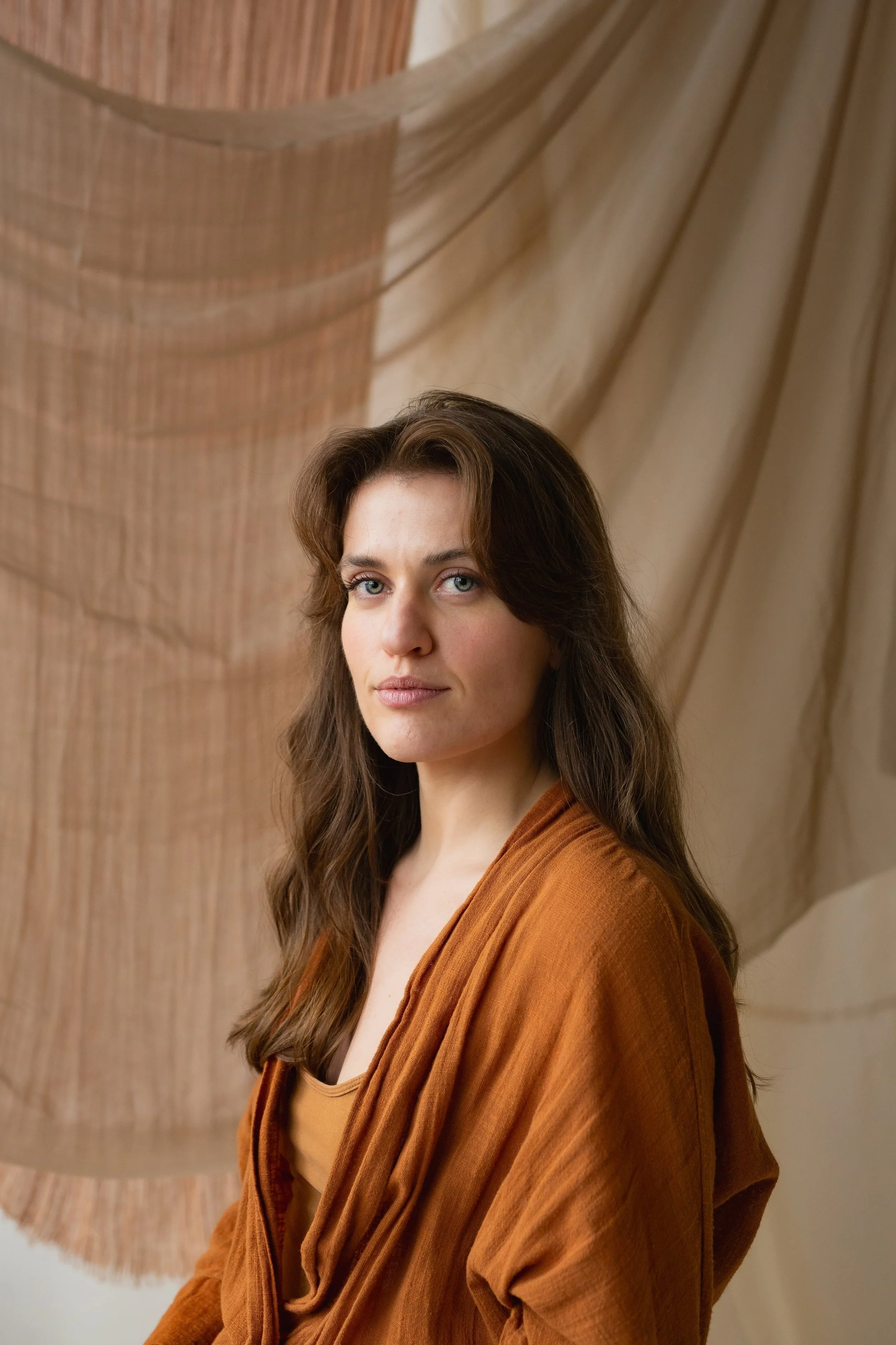 A woman with long brown hair and light skin wearing a rust-colored shirt, standing in front of a large wooden wall sculpture and beige draped fabric.