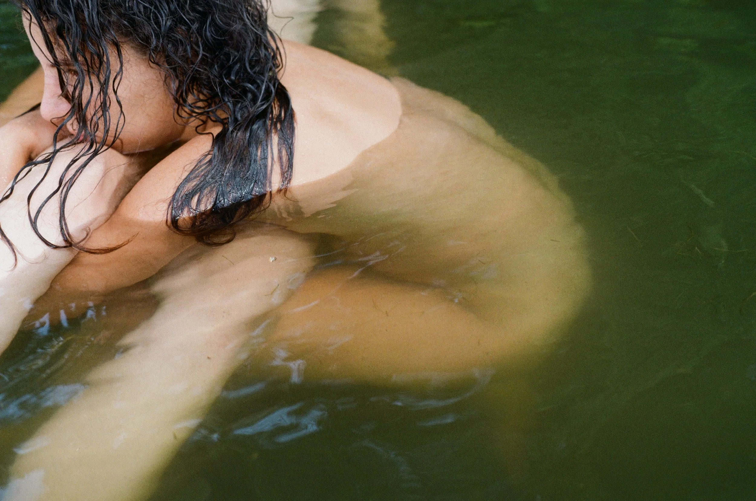 A person with curly dark hair partially submerged in water, resting their head on their arms in a natural, outdoor setting.