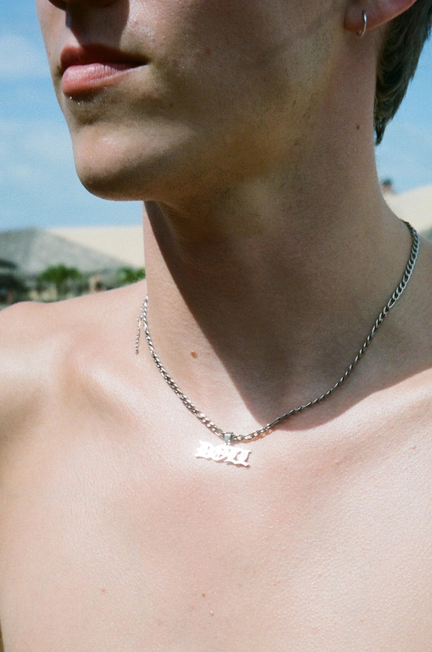 Close-up of a person's neck and chest with a silver chain necklace that has a pendant spelling 'BABY' and small earrings, with blue sky and blurred background.