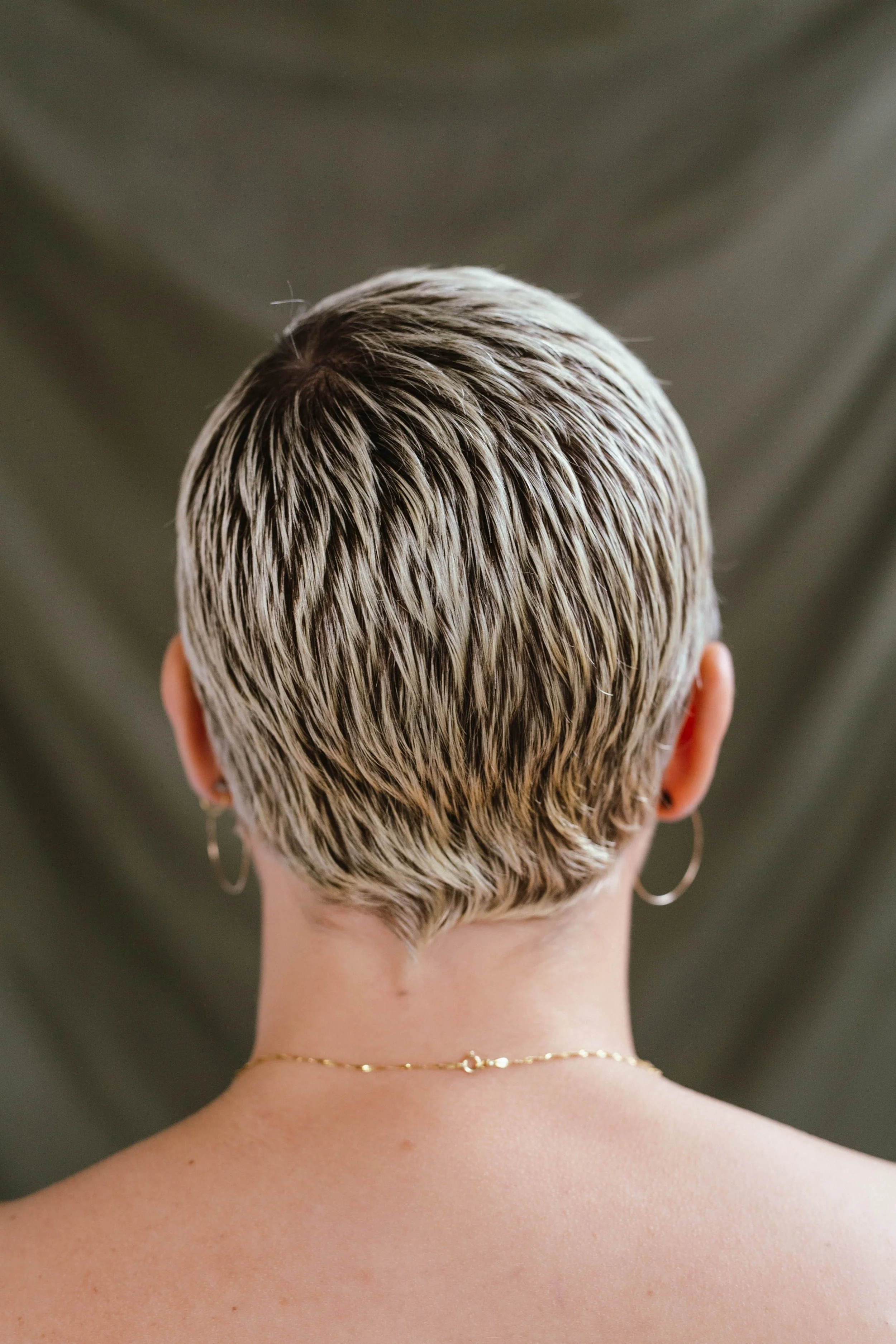 Back of a person's head with short, textured, blonde-tipped hair, wearing hoop earrings and a delicate gold chain necklace, with skin showing on the neck and shoulders, against a blurred neutral background.