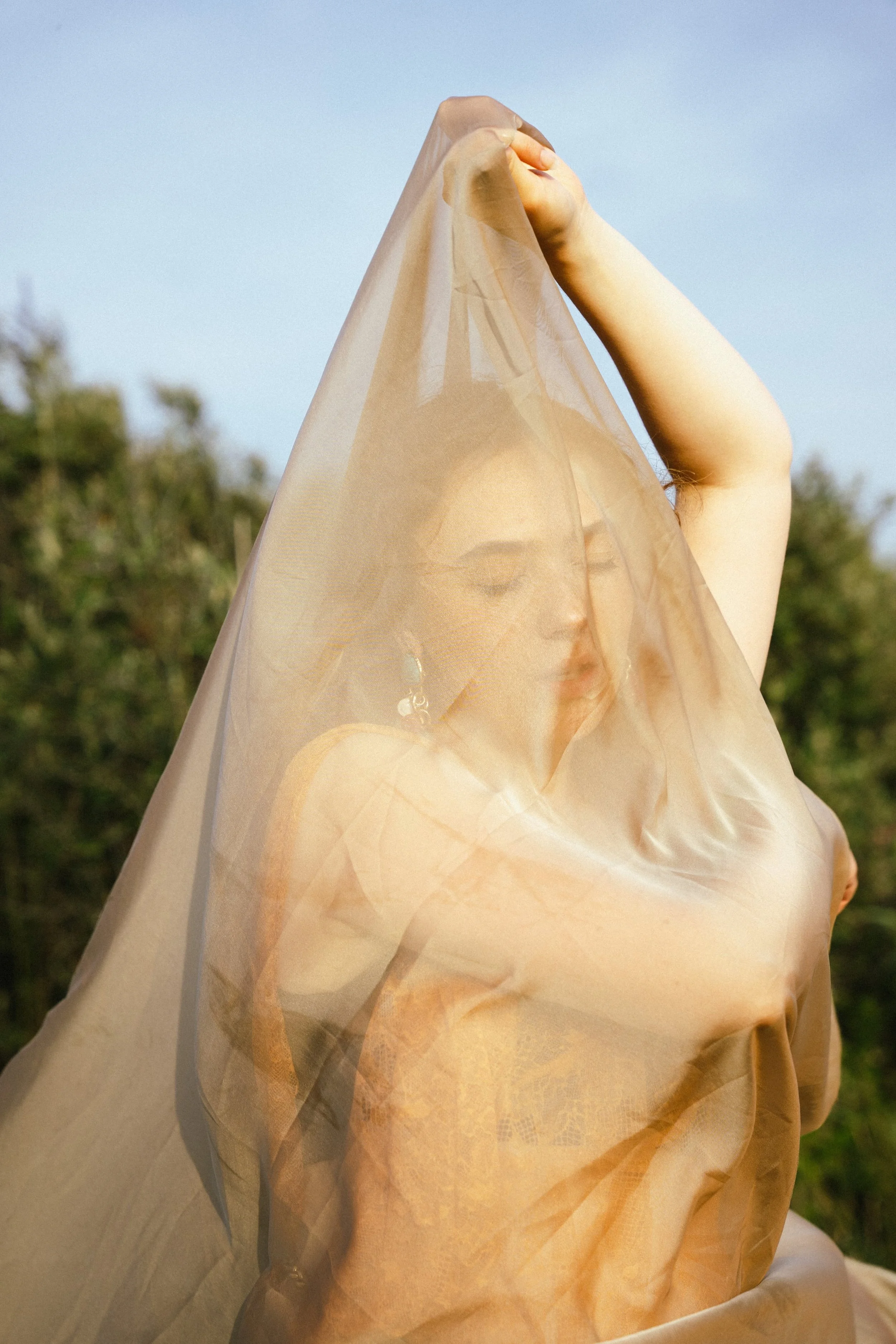 A woman with dark hair and hoop earrings holding a semi-transparent beige fabric over her face and head outdoors with a blurred background of trees and blue sky.