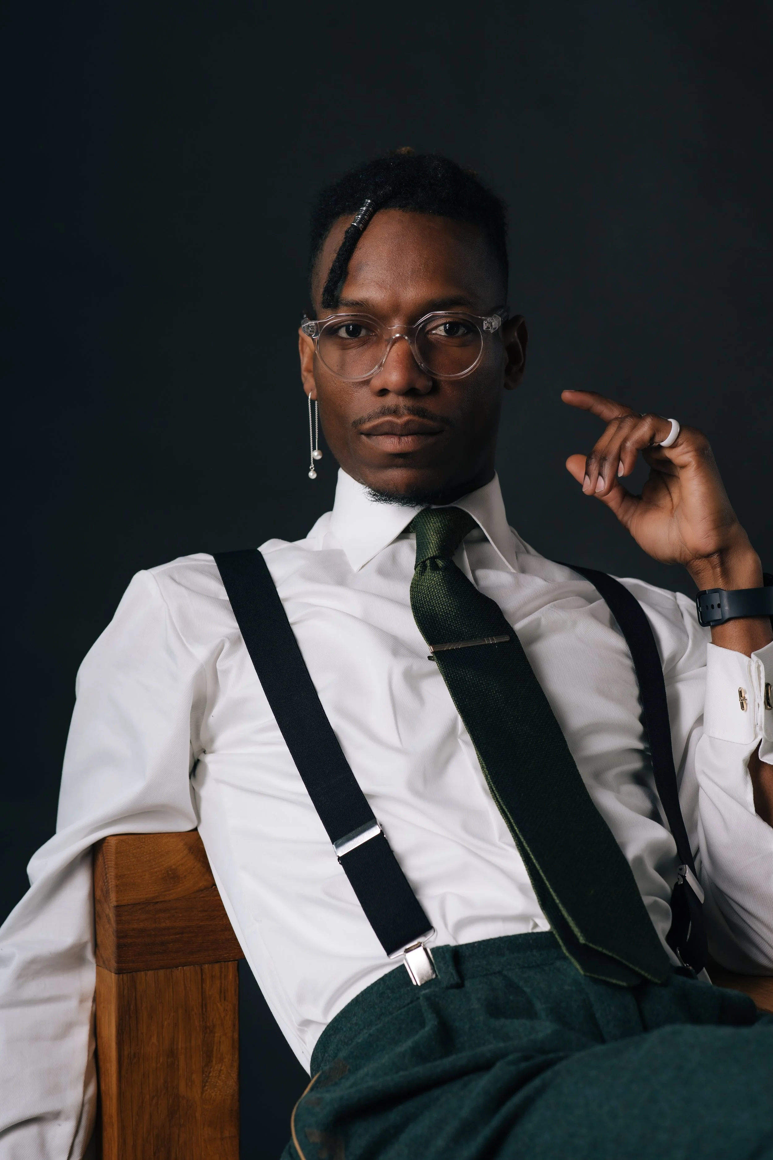 A man wearing clear glasses, a white shirt, a green tie, and suspenders, sitting on a wooden chair against a dark background, with one arm raised and pointing to his temple.