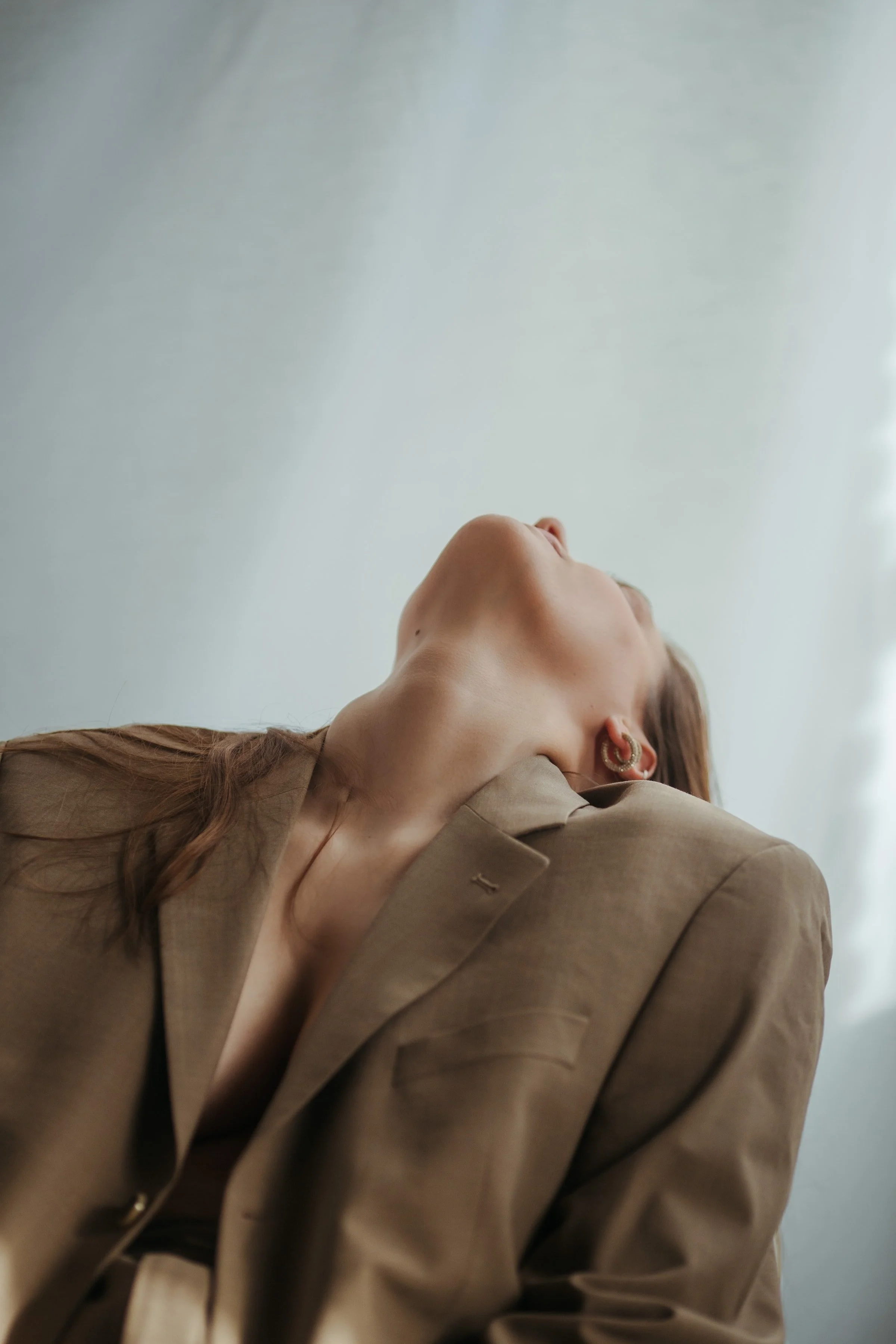 A woman with light skin and brown hair looking upward, wearing gold hoop earrings and a beige blazer.