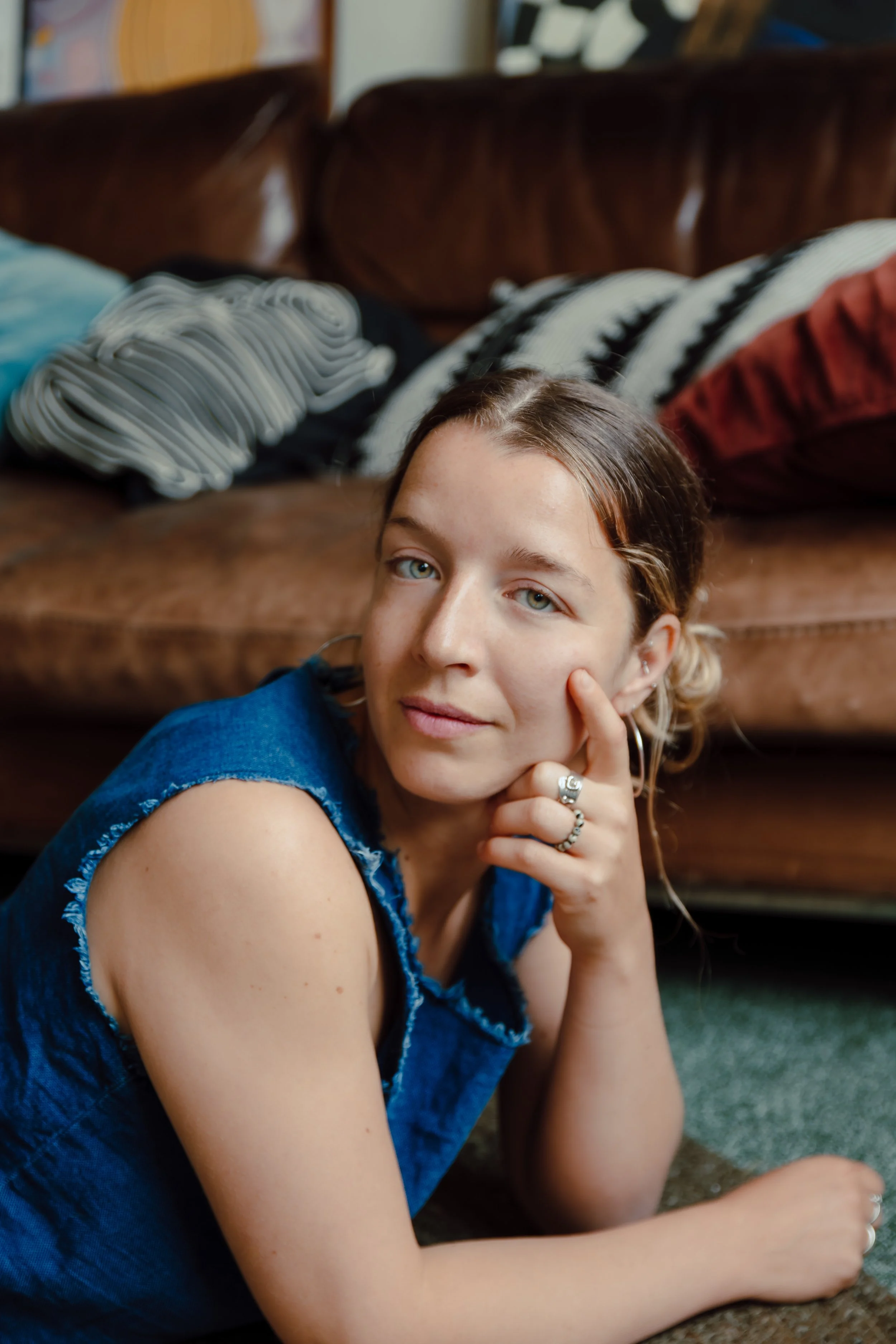 Close-up of a young woman with blonde hair and blue eyes, resting her head on her hand and looking at the camera, sitting on the floor near a brown leather couch with colorful throw pillows.