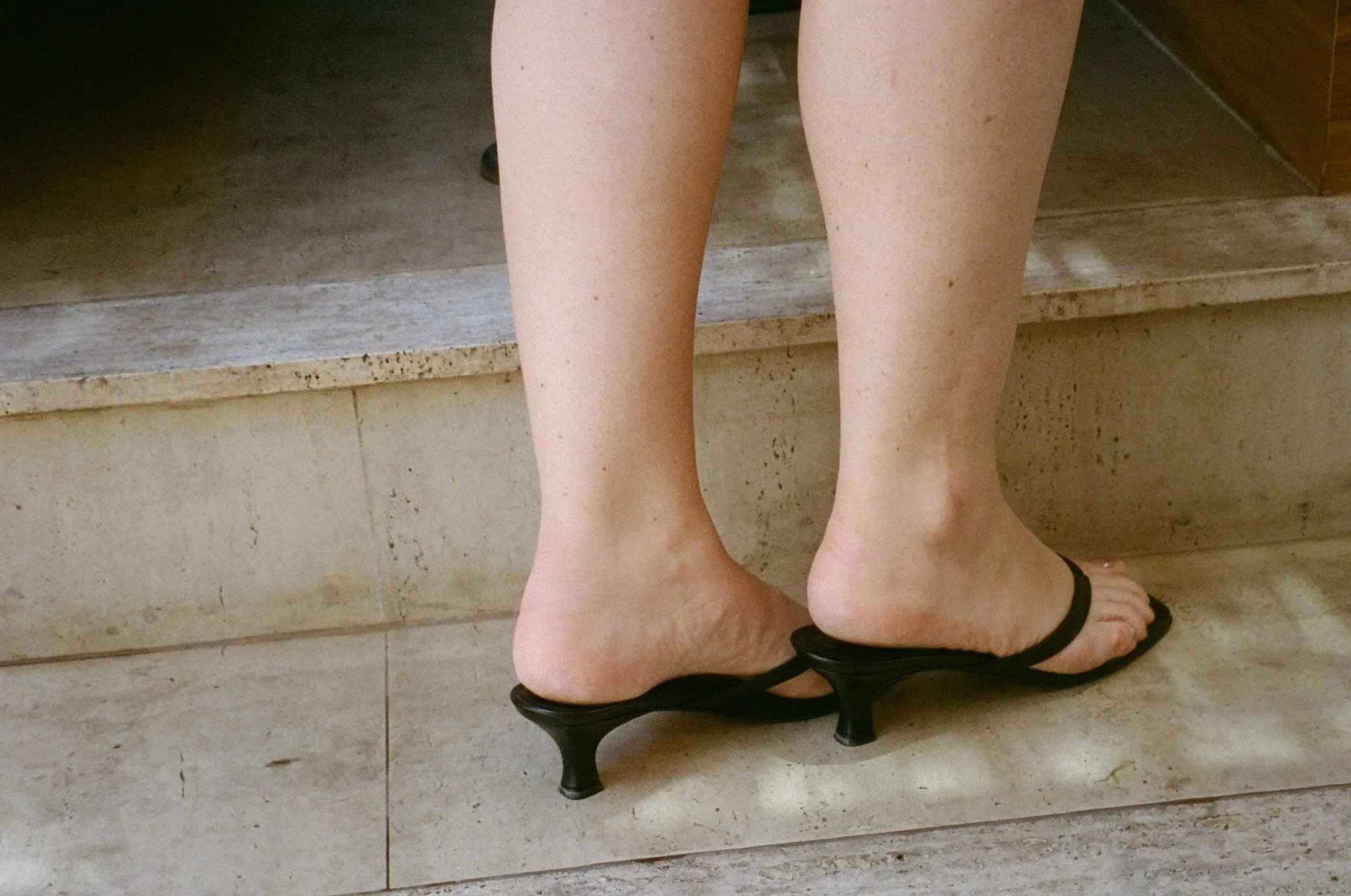 Close-up of a person's bare legs and feet, wearing black sandals, standing on a tiled floor next to a set of stairs.