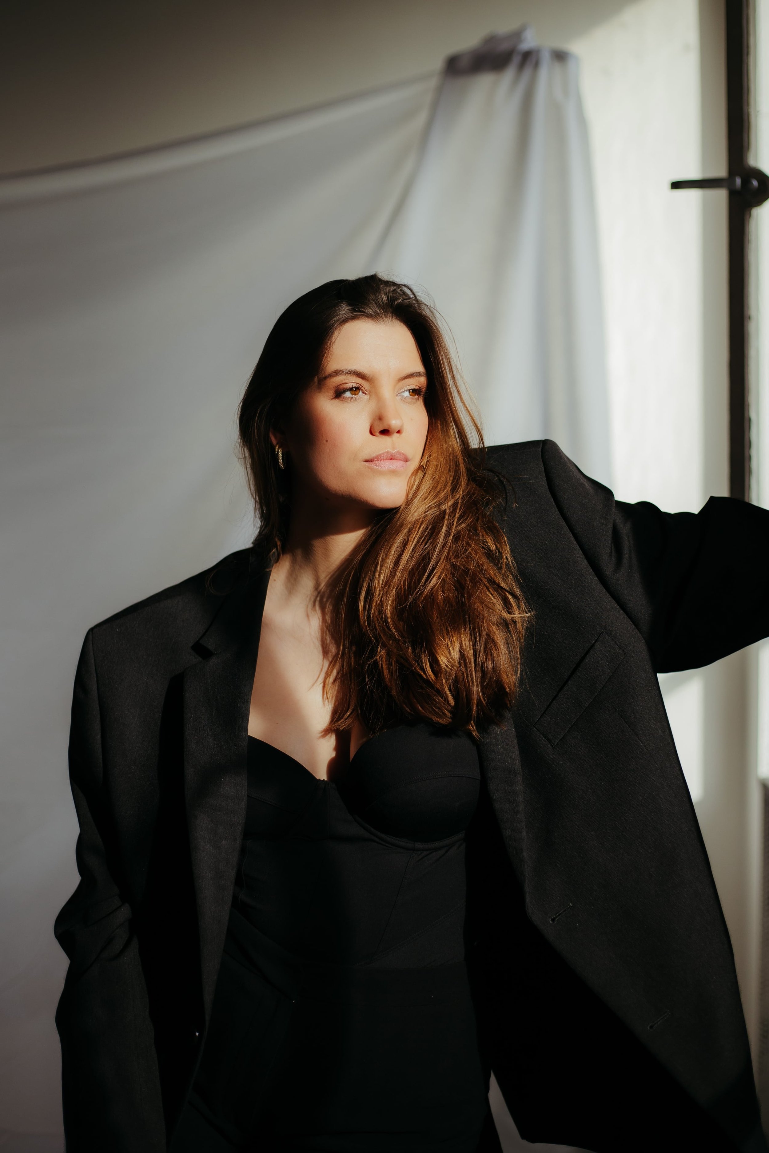 A woman with long brown hair wearing a black dress and a black blazer, sitting in front of a light-colored backdrop near a window with natural light, looking off to the side.