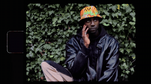 Person wearing a colorful bucket hat and black jacket, sitting in front of green ivy, resting their face on their hand.