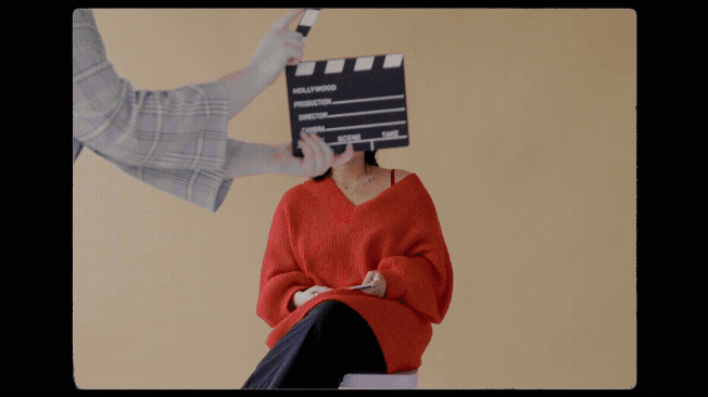 Person wearing an orange sweater sitting on a chair with their face covered by a film slate held by another person in a checkered shirt.
