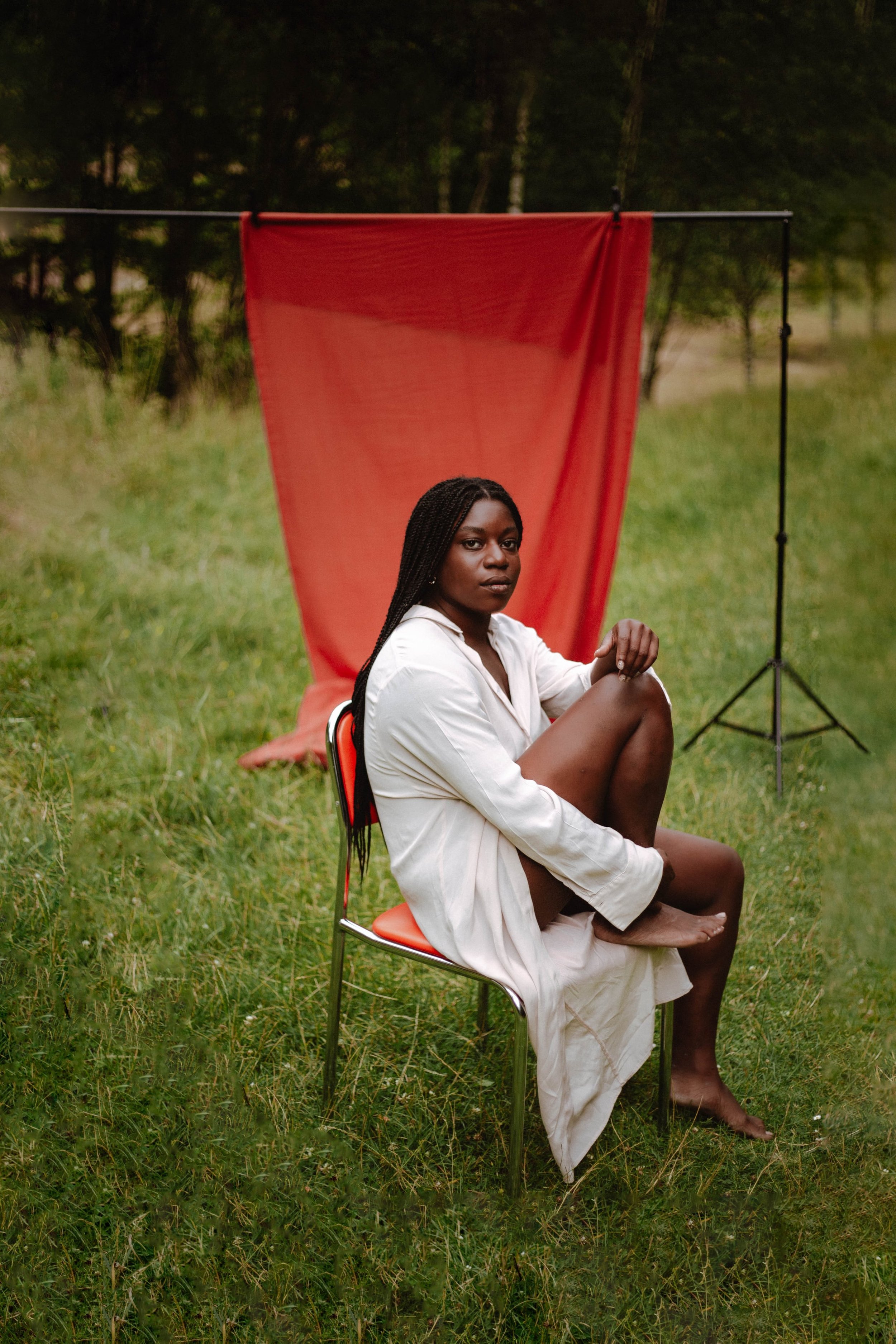 A woman with braided hair sitting barefoot on a red chair in a grassy outdoor area, with a red fabric hanging on a stand behind her and trees in the background.