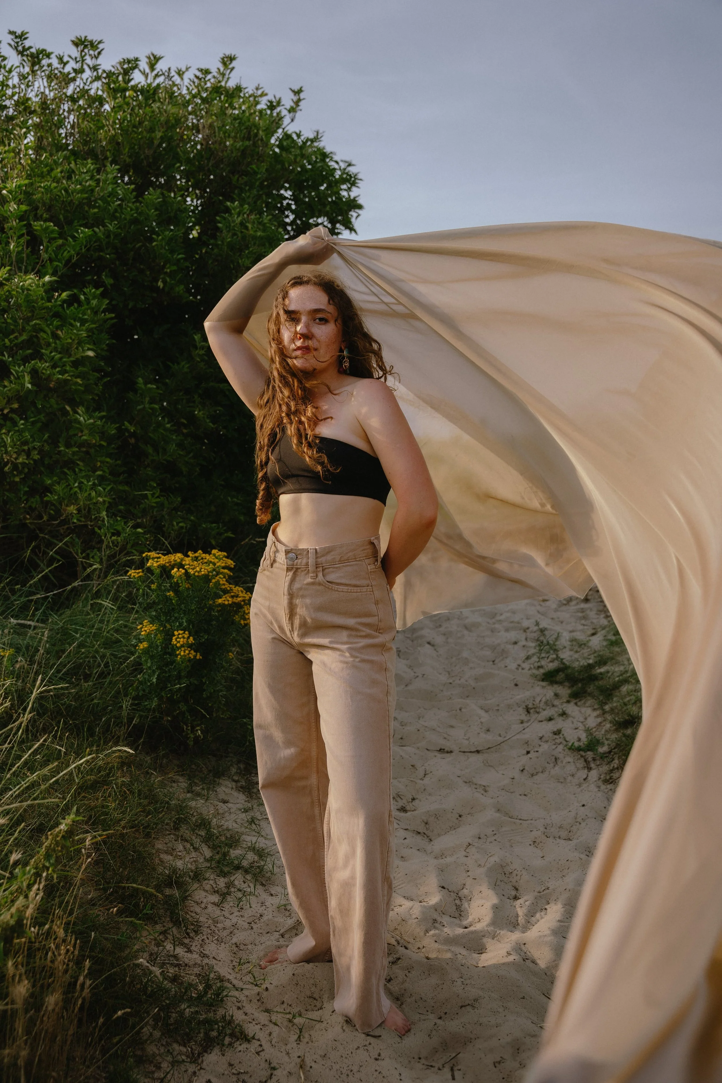 A woman with curly hair stands on a sandy path beside green bushes, holding a beige fabric backdrop. She is wearing a black strapless top and beige high-waisted pants, looking at the camera with a serious expression.