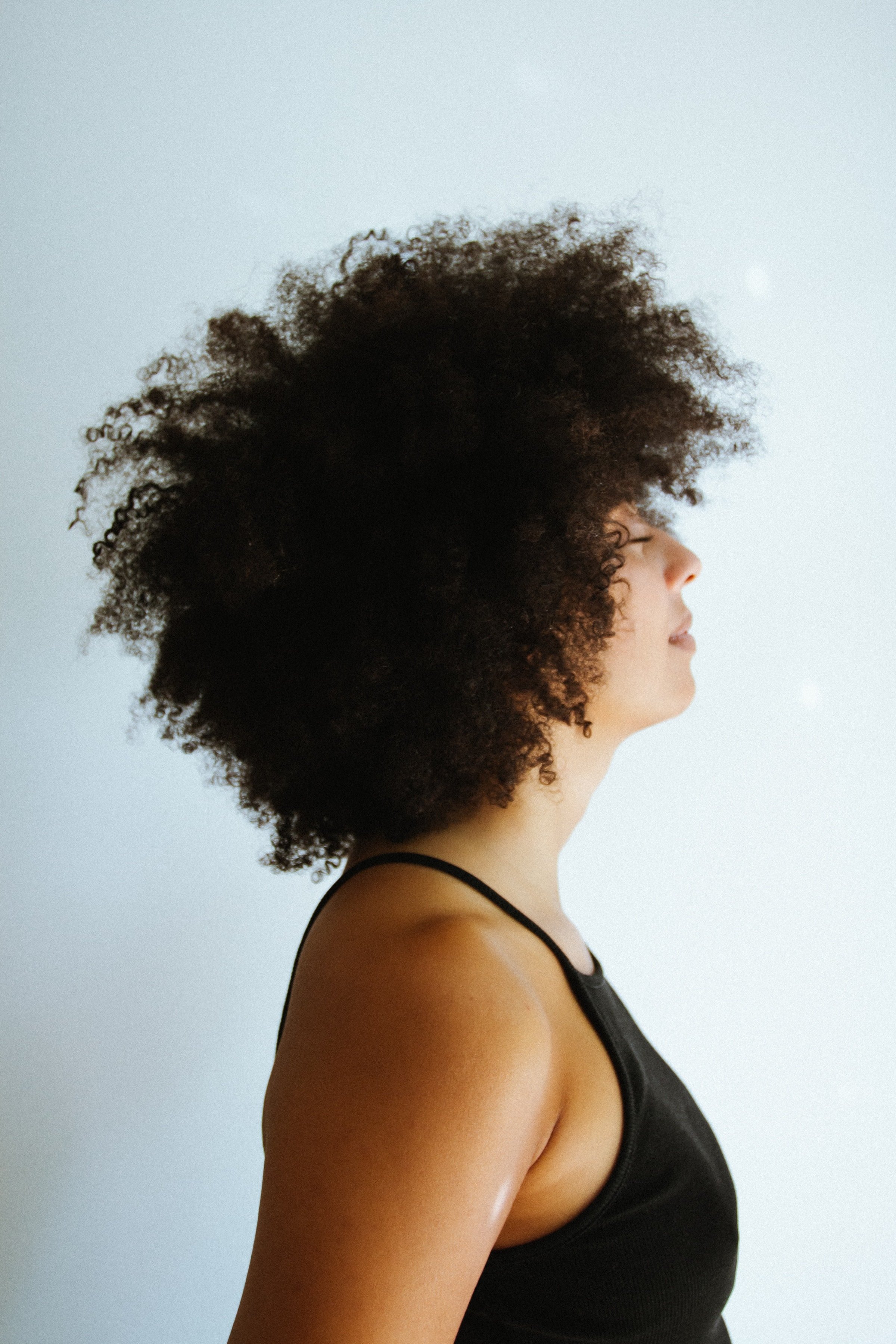 Profile of a woman with curly afro hairstyle, wearing a black tank top, facing sideways against a plain light-colored background.