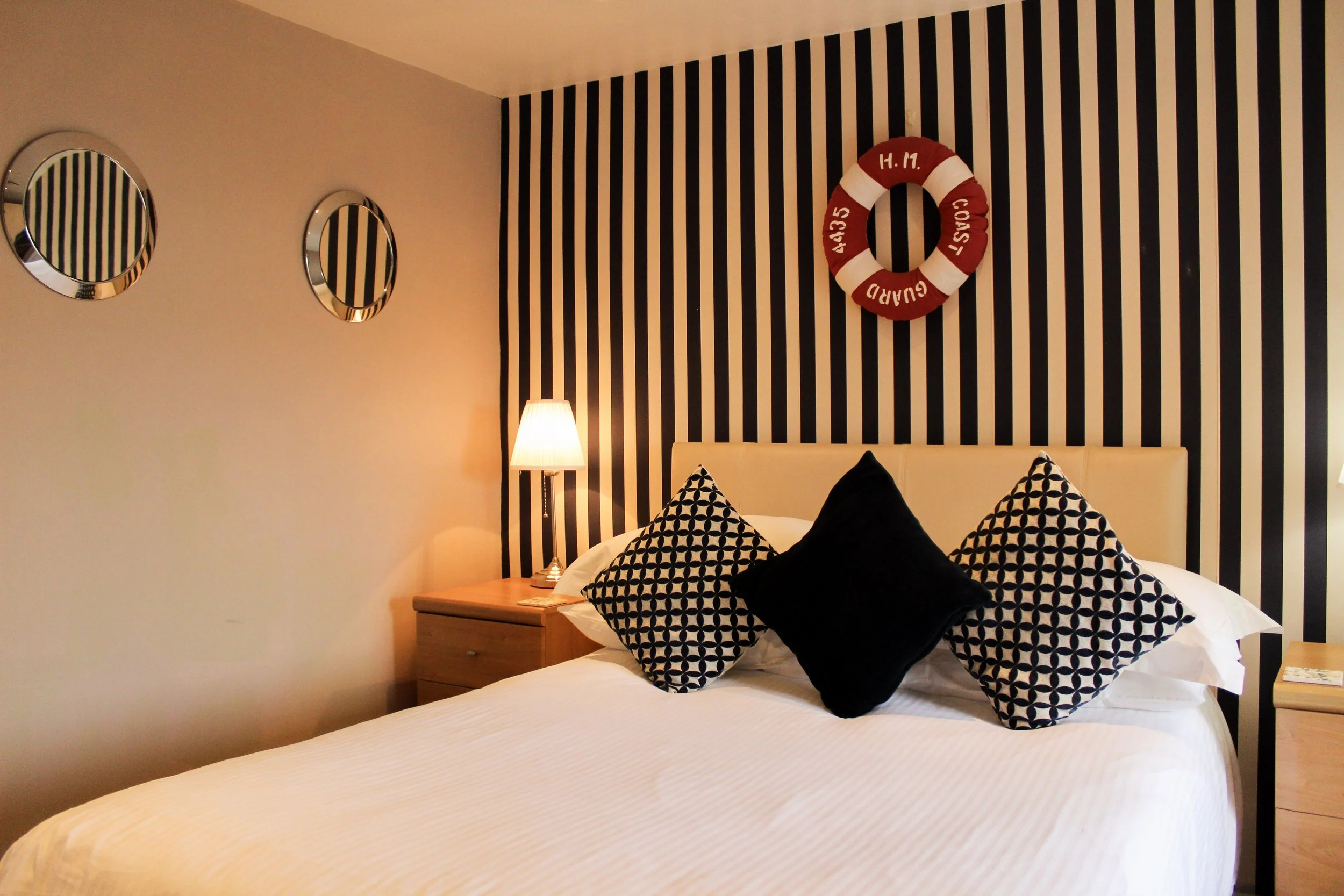 Side image of double bed with three pillows and stripe wallpaper with a life ring on the wall.