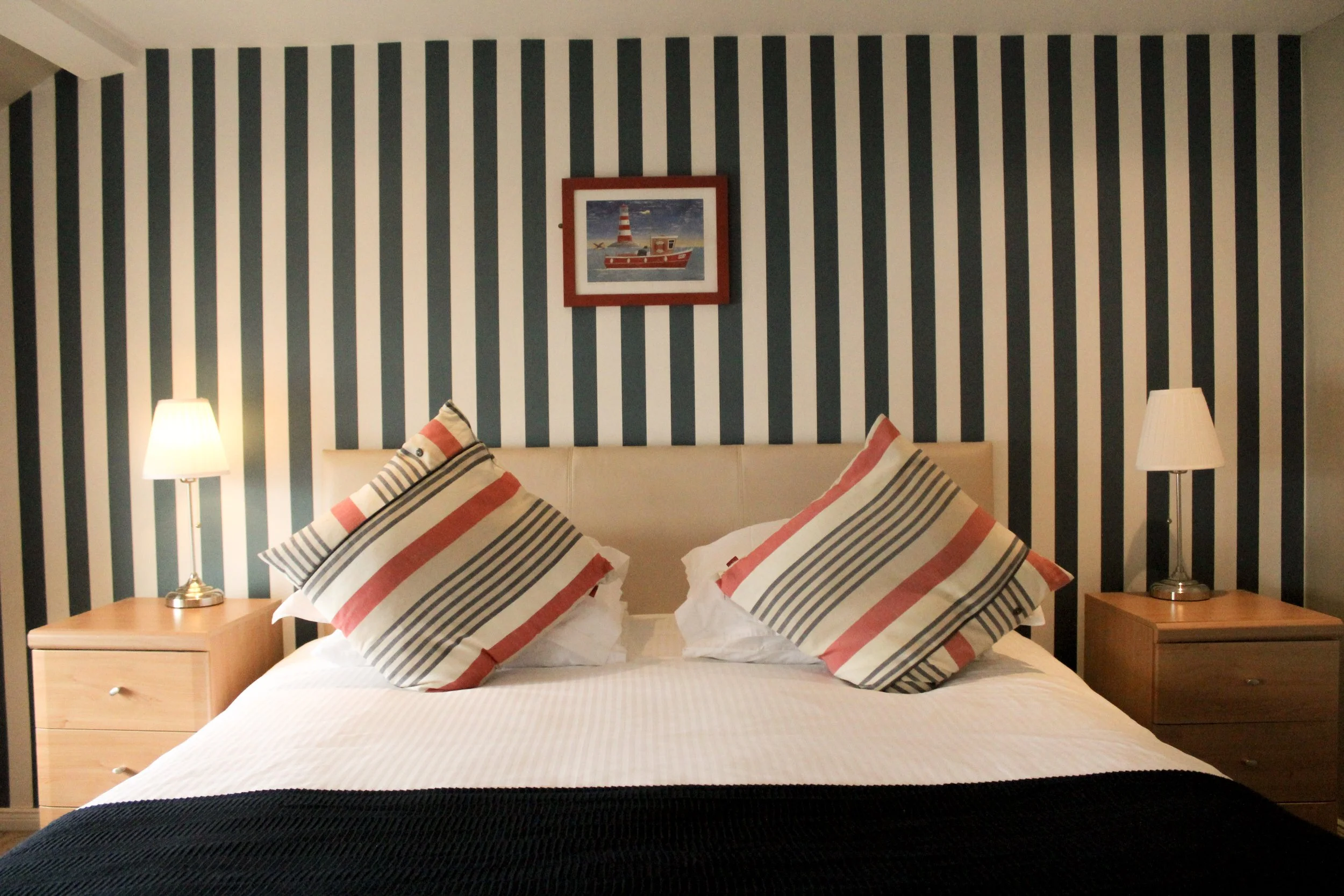 Image of Pier 36 guest house room with bed and stripe wallpaper. Bedside tables on each side with a single lamp.