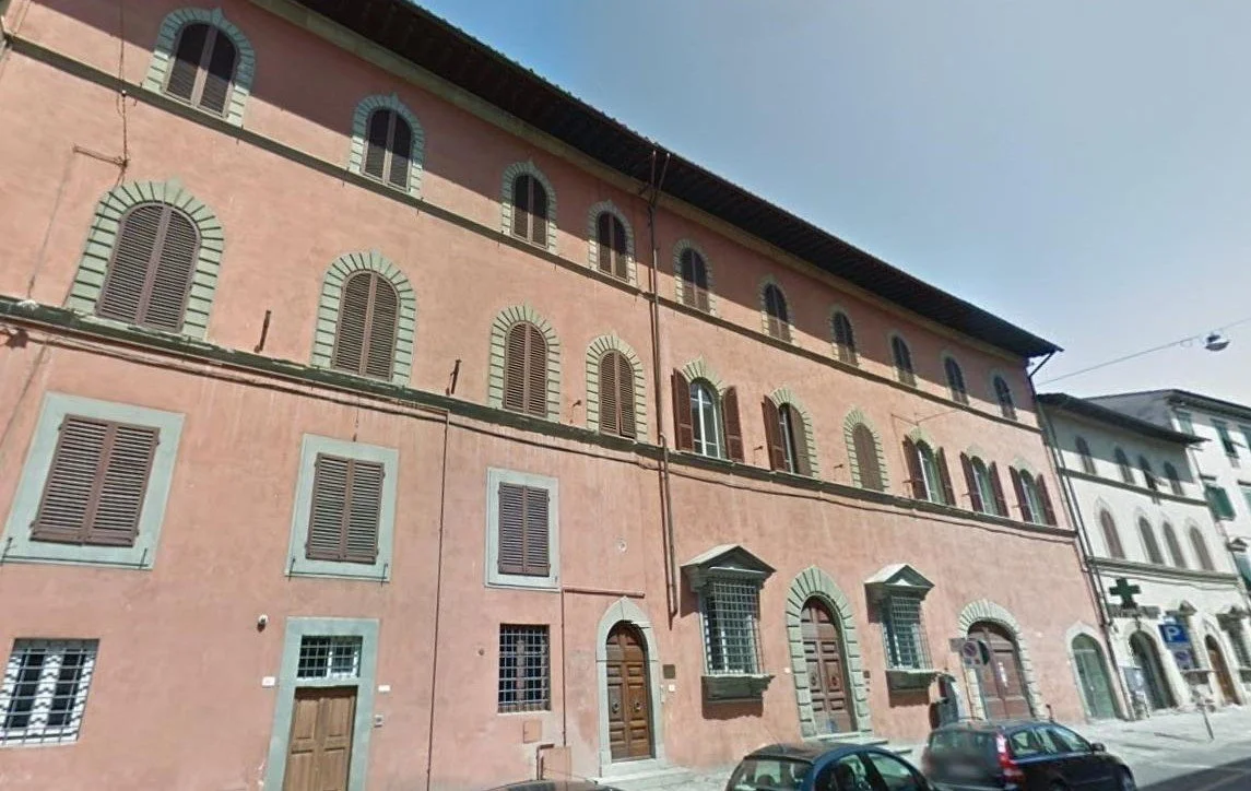 Edificio storico di colore rosa con finestre ad arco e persiane marroni, situato in una strada urbana con auto parcheggiate davanti.