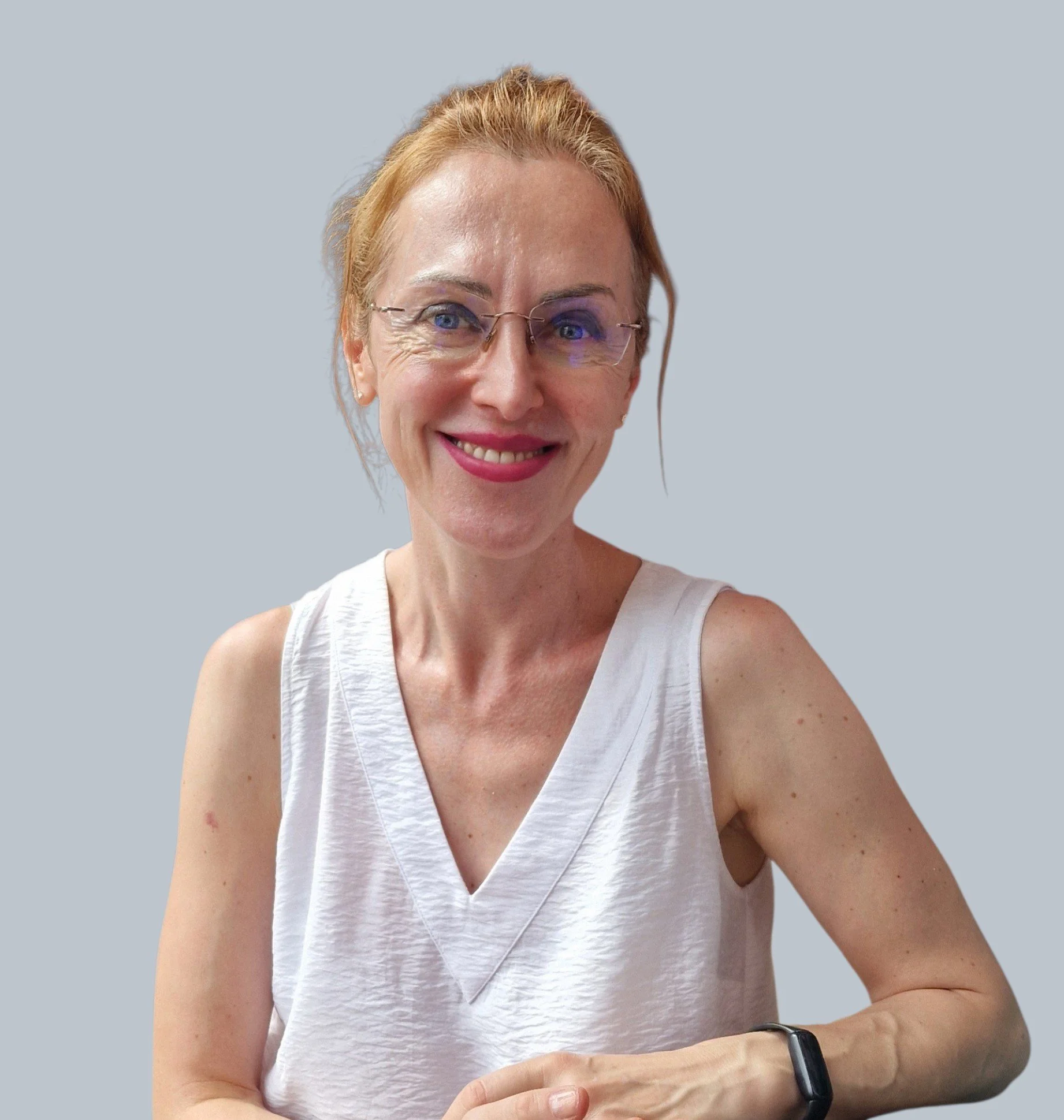 A woman with red hair and glasses smiling at the camera, wearing a white sleeveless top and a black smartwatch on her left wrist. She is in front of a light gray background.