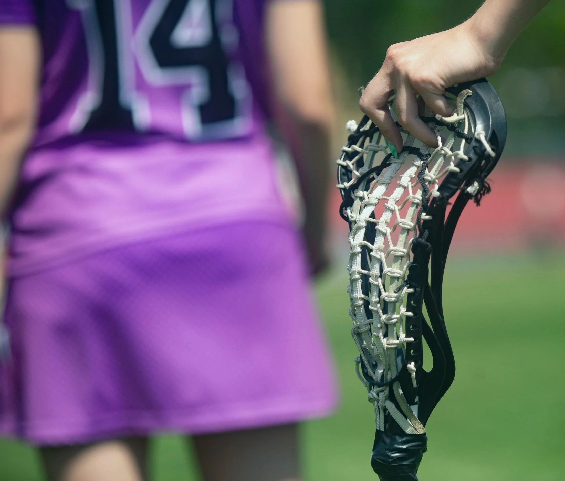 Person holding a lacrosse stick on a field, with another person in a purple uniform nearby.