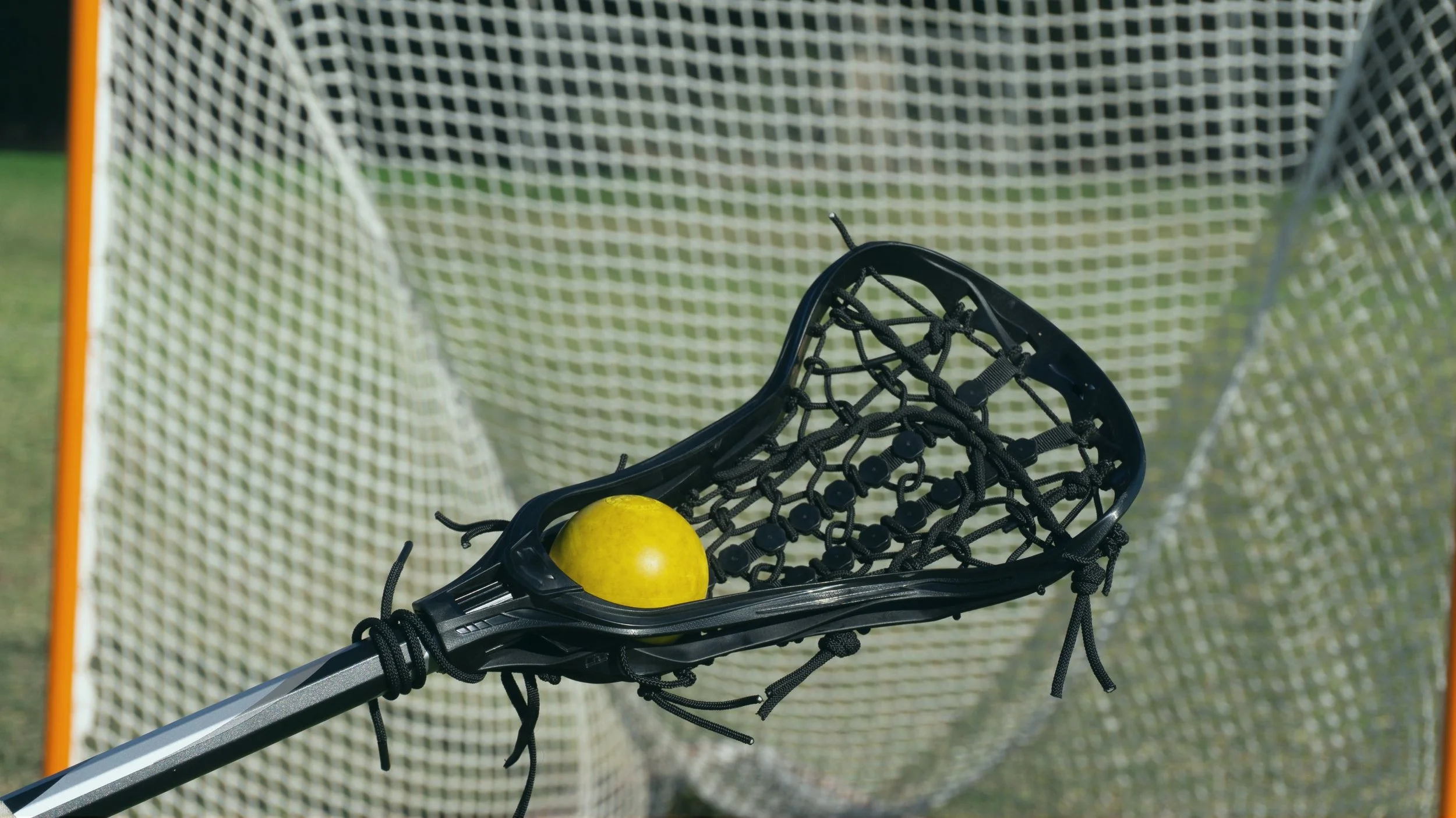 Lacrosse stick with a yellow ball inside the net, positioned in front of a lacrosse goal net.