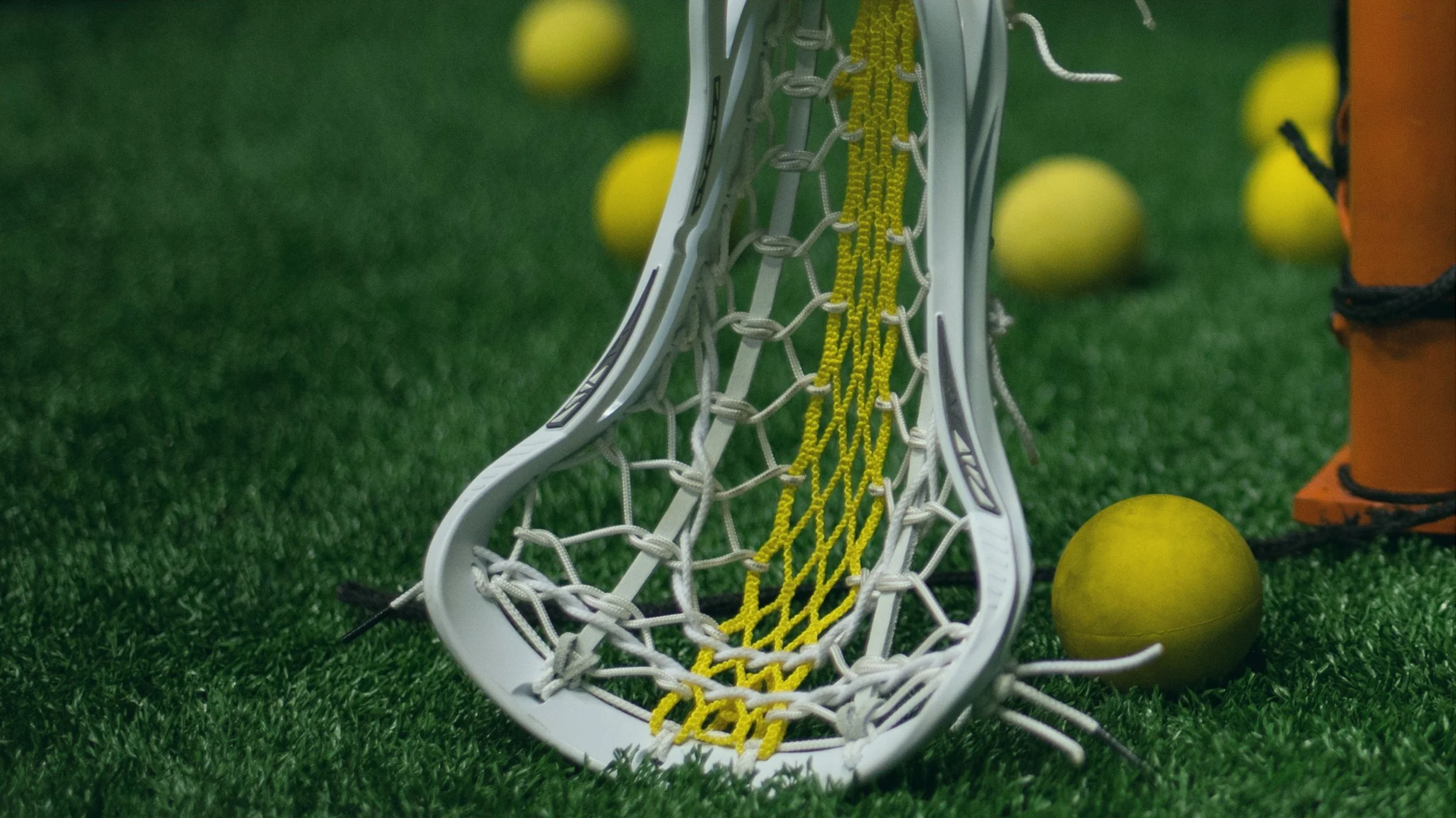 A lacrosse stick with a white frame and yellow netting resting on artificial green turf surrounded by yellow lacrosse balls and a section of orange lacrosse goal post.