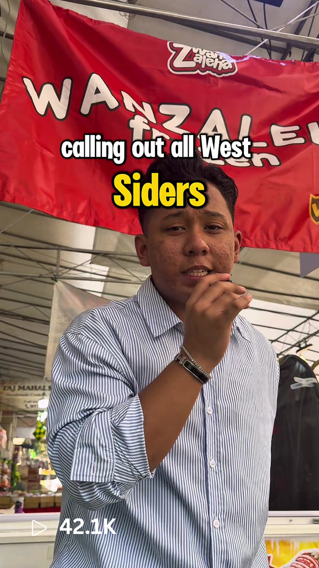 A young man standing in front of a red banner that reads 'Wazala Zapata' with a backdrop of stalls at an outdoor market.