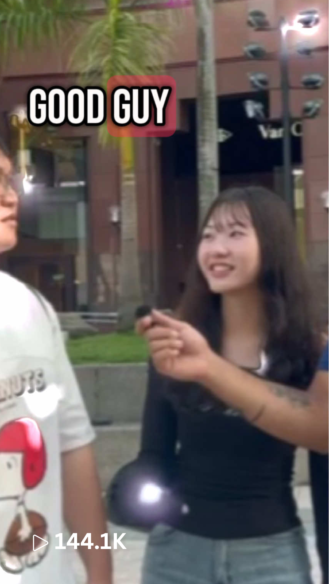 A woman smiling and holding a microphone in front of a man. There is a sign that says 'Good Guy' above them, and they are outdoors in front of a building with trees.