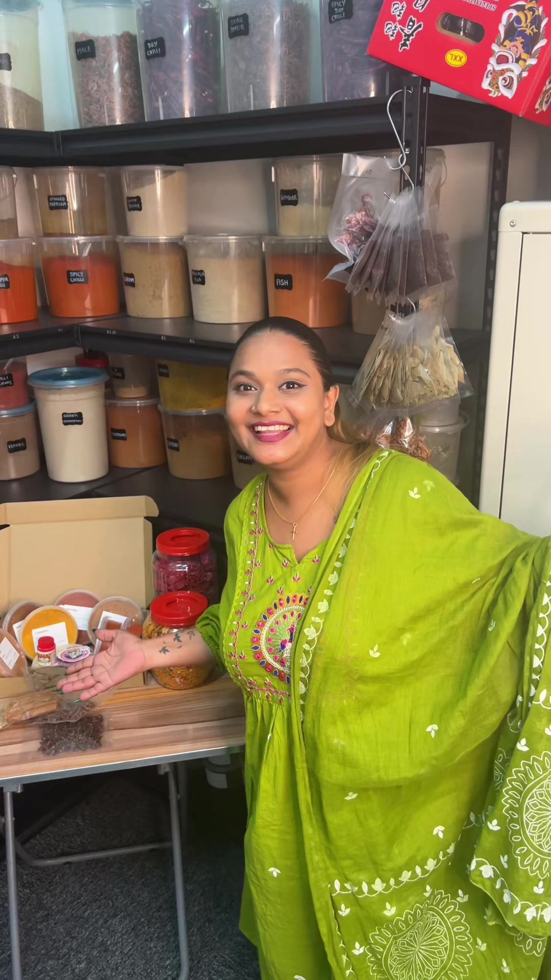 Woman smiling and gesturing with her hands in front of shelves filled with various spices and herbs, with plastic bags of dried herbs hanging nearby, in a kitchen or spice store.