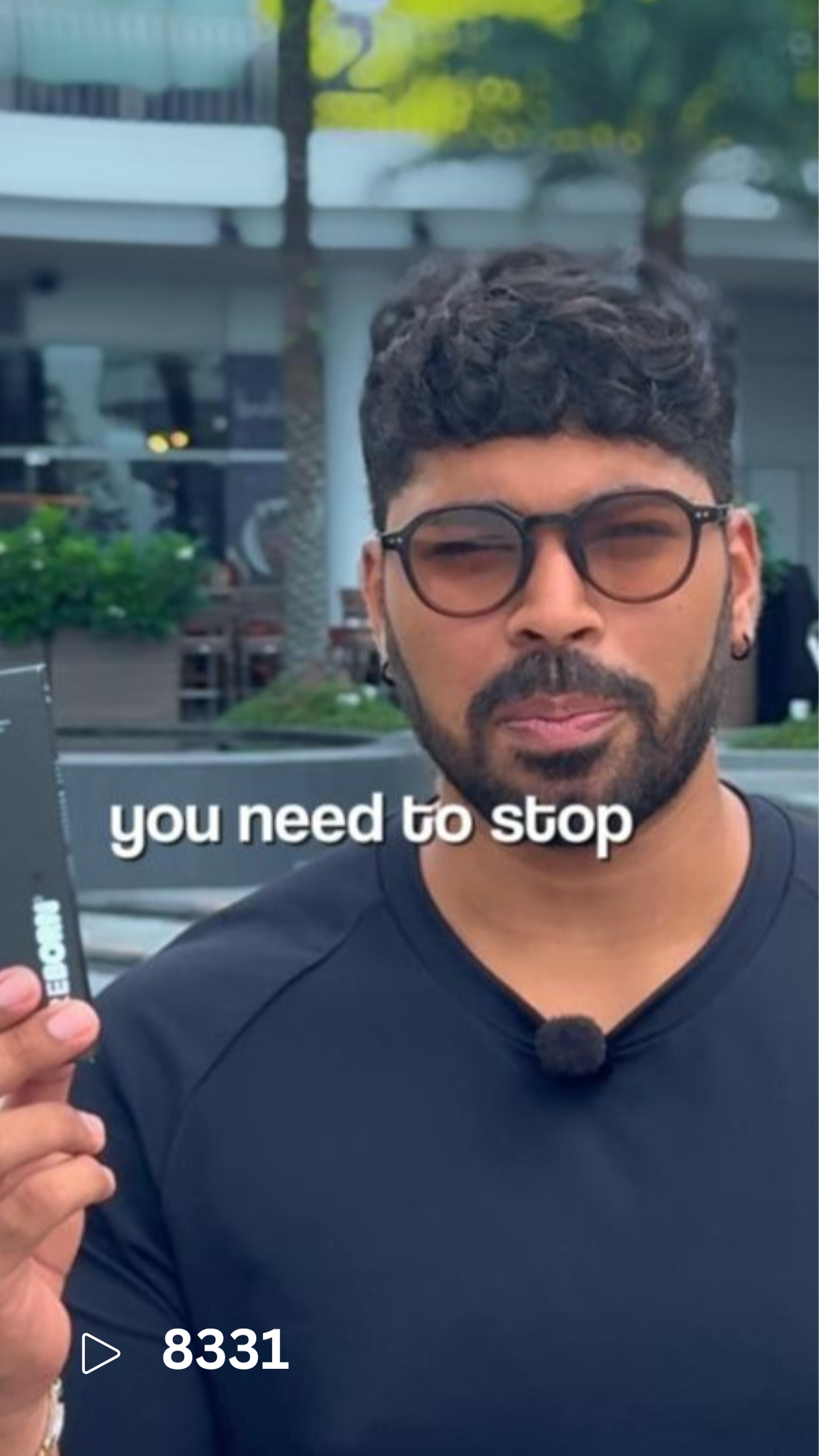 A man with curly hair, glasses, and a beard holding a device with a serious expression, outdoors with buildings and trees in the background.