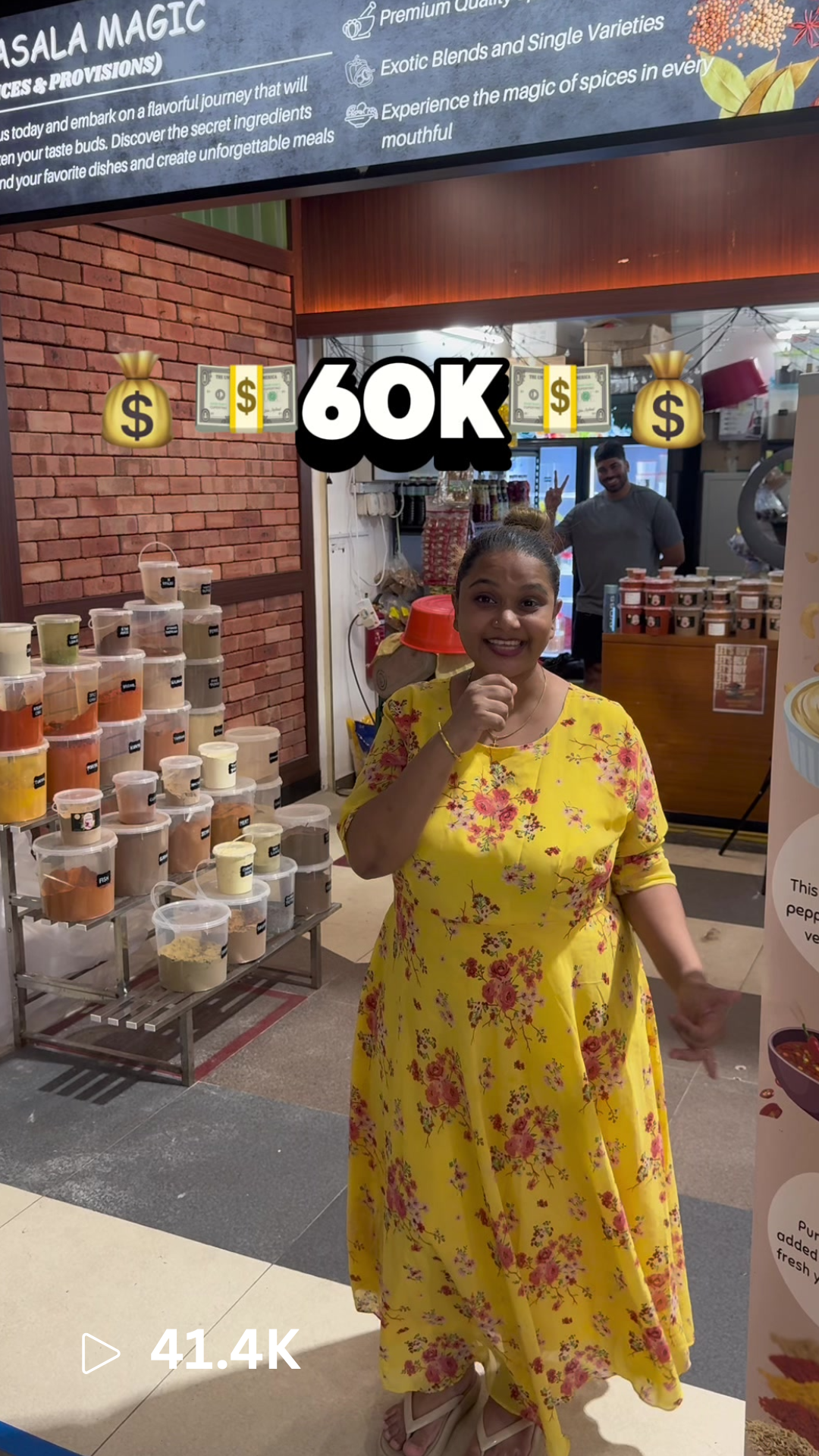 A woman in a yellow floral dress standing inside a store with various colorful powders in transparent containers behind her, and a man in a gray T-shirt in the background making a peace sign.