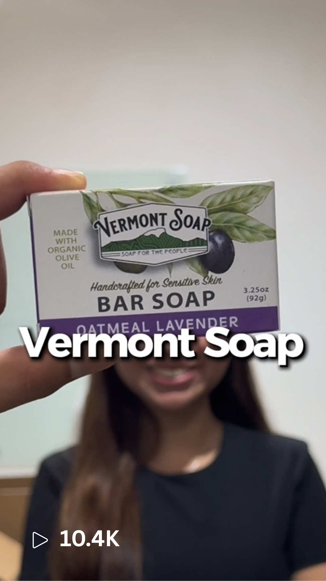 Person holding a Vermont Soap oatmeal lavender bar soap box in front of their face, smiling, with a beige wall background.