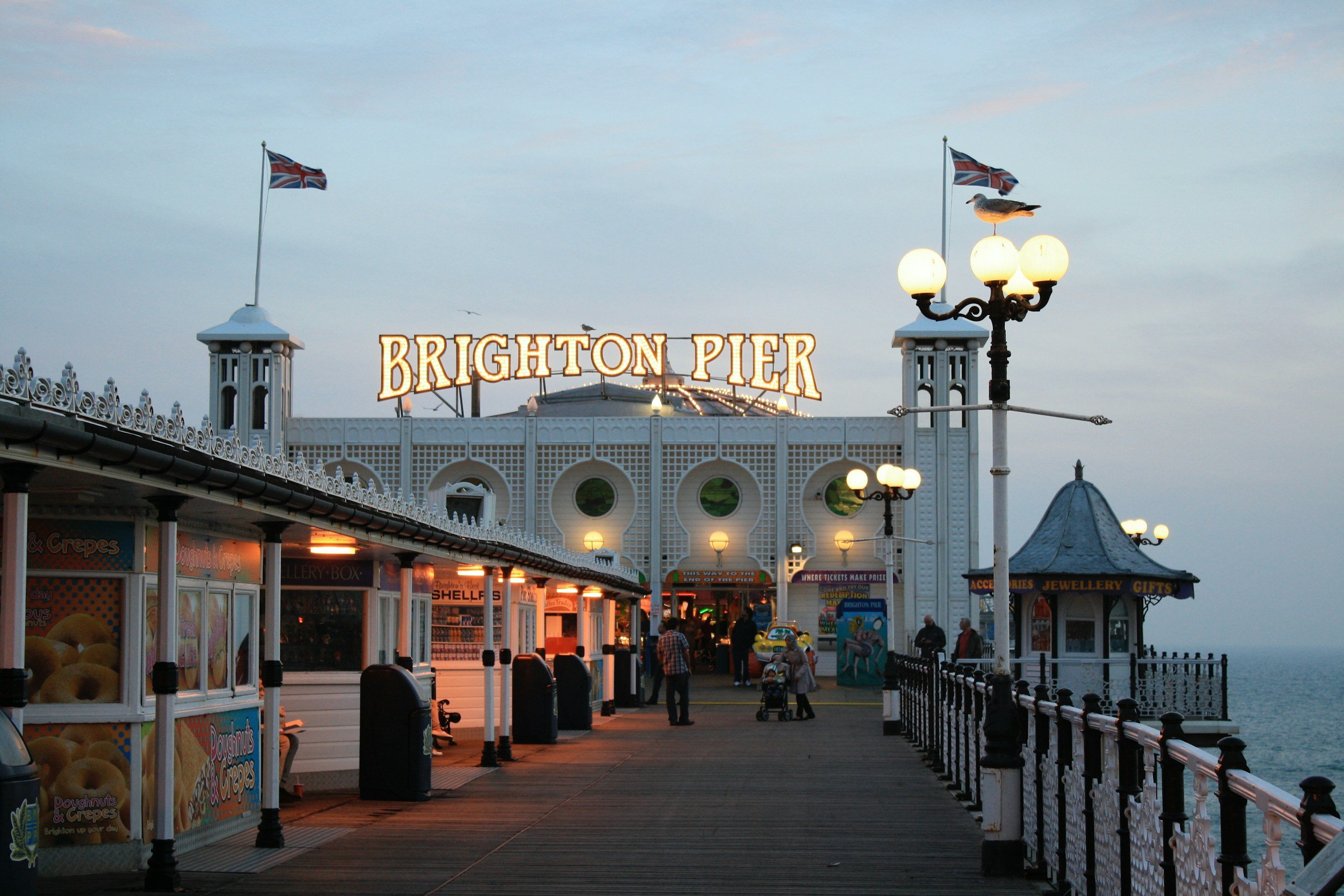 Brighton Pier at dusk with the sign lit up, flags, seagulls, and street lamps. Visitors walk along the pier with shops and vendors, and the ocean is visible to the right.