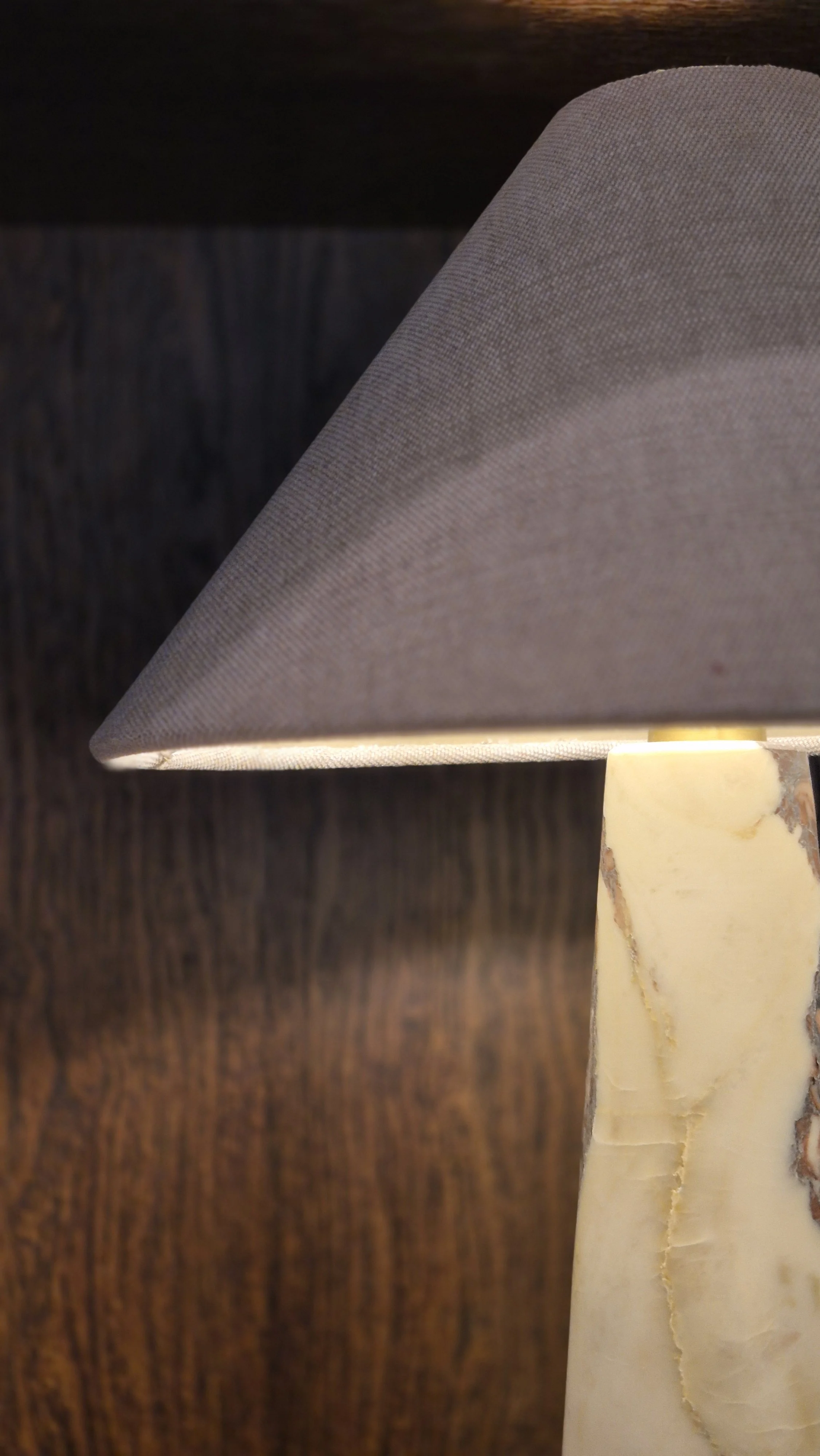 Close-up of a table lamp with a beige marble base and a grey fabric lampshade, against a dark wooden background.