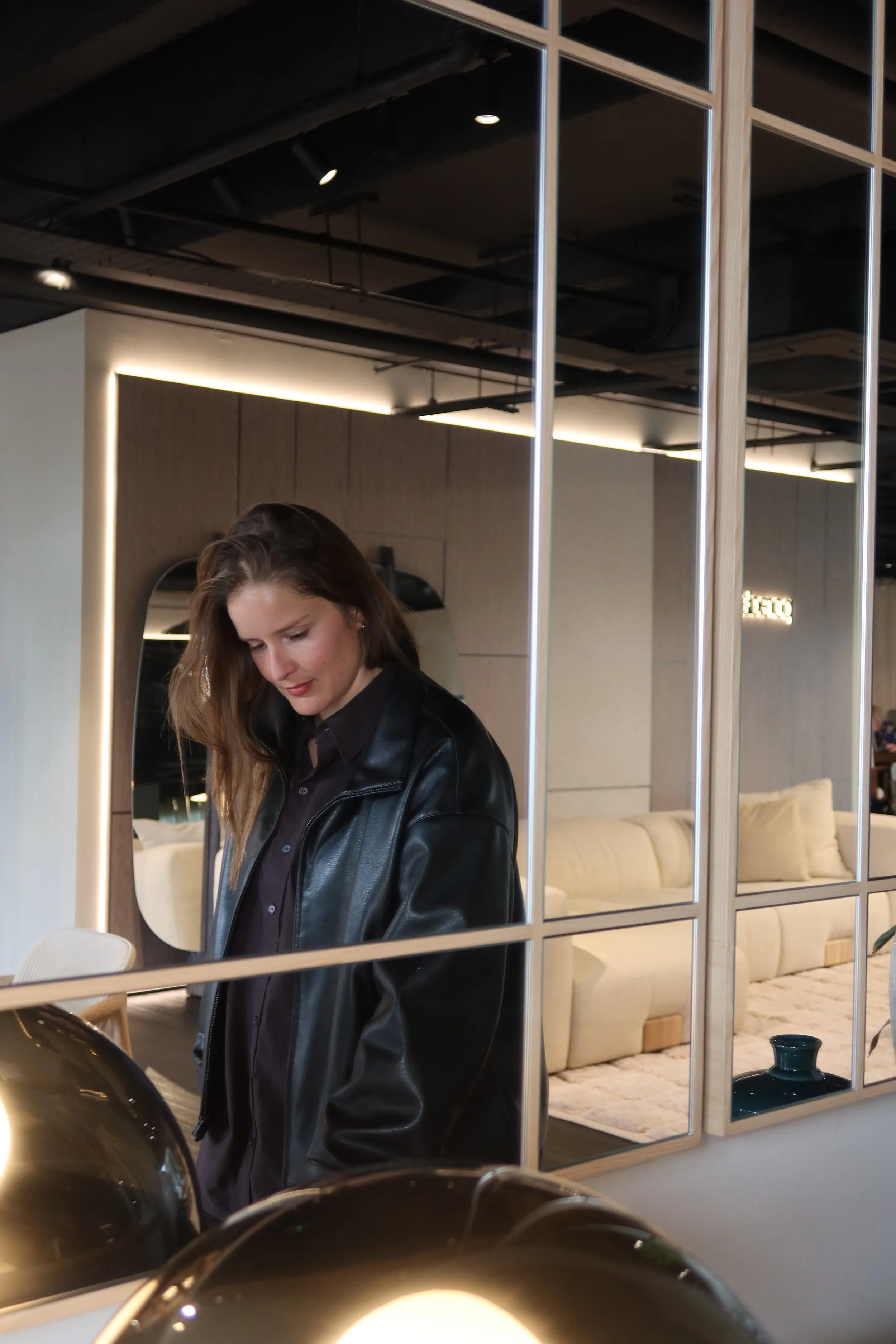 A woman with long brown hair wears a black leather jacket and black shirt, looking down, in front of a mirror in a modern, well-lit room with beige couch and wooden wall panels.