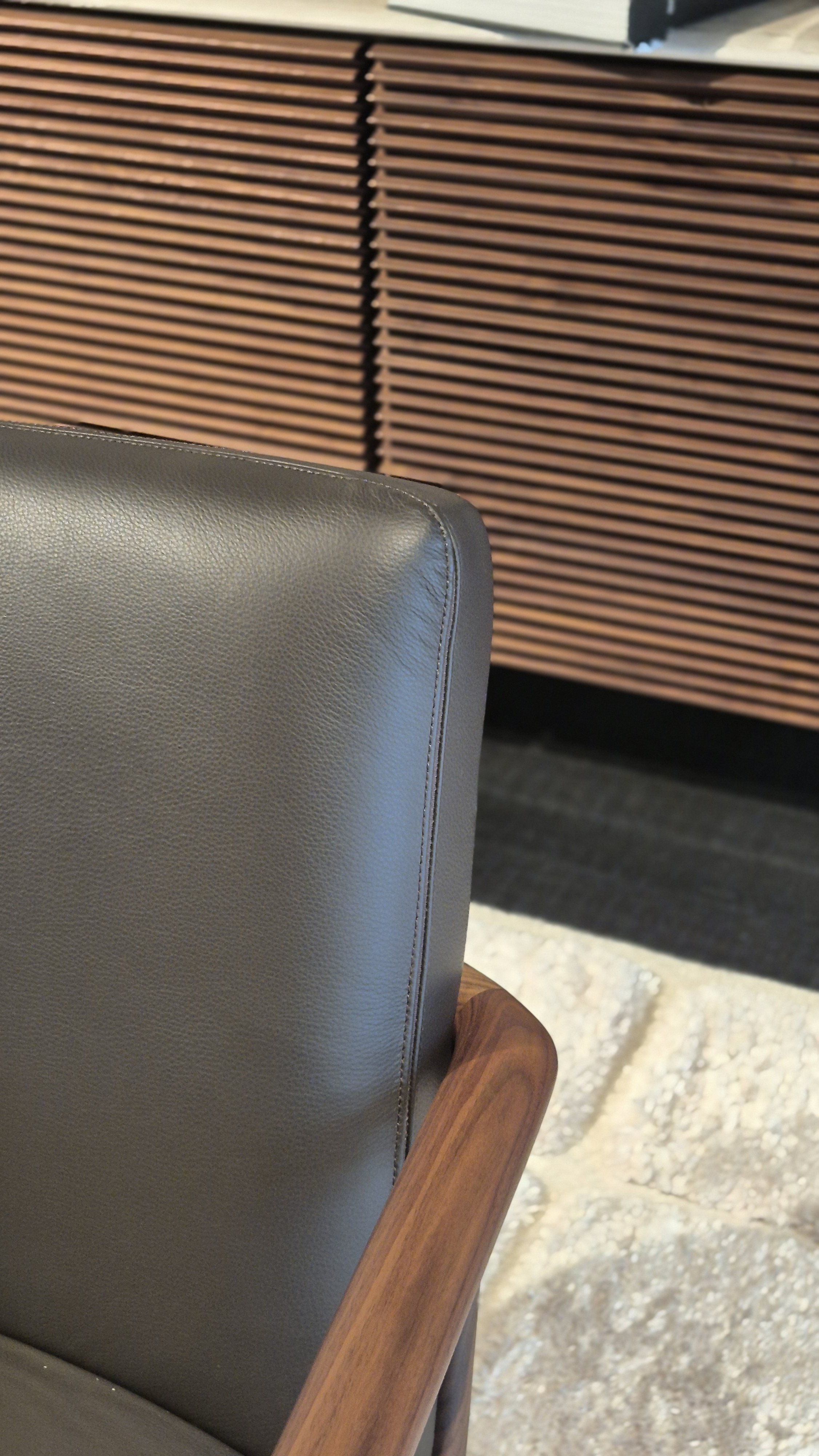 Close-up of a gray upholstered chair with wooden armrests, with a textured stone floor and wooden slatted furniture in the background.