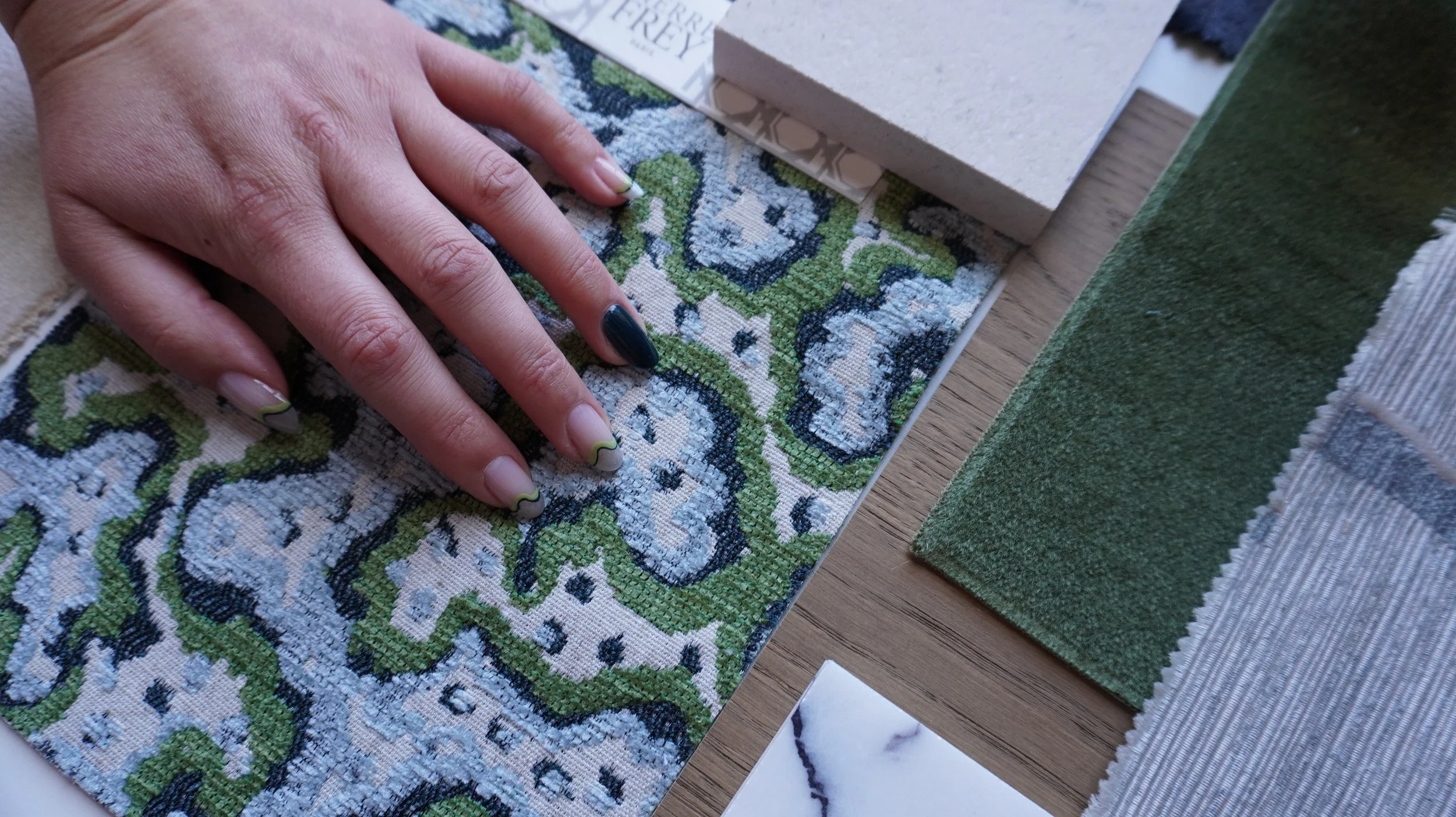 A hand with manicured nails resting on a patterned fabric sample. There are various fabric samples and color swatches nearby on a table.