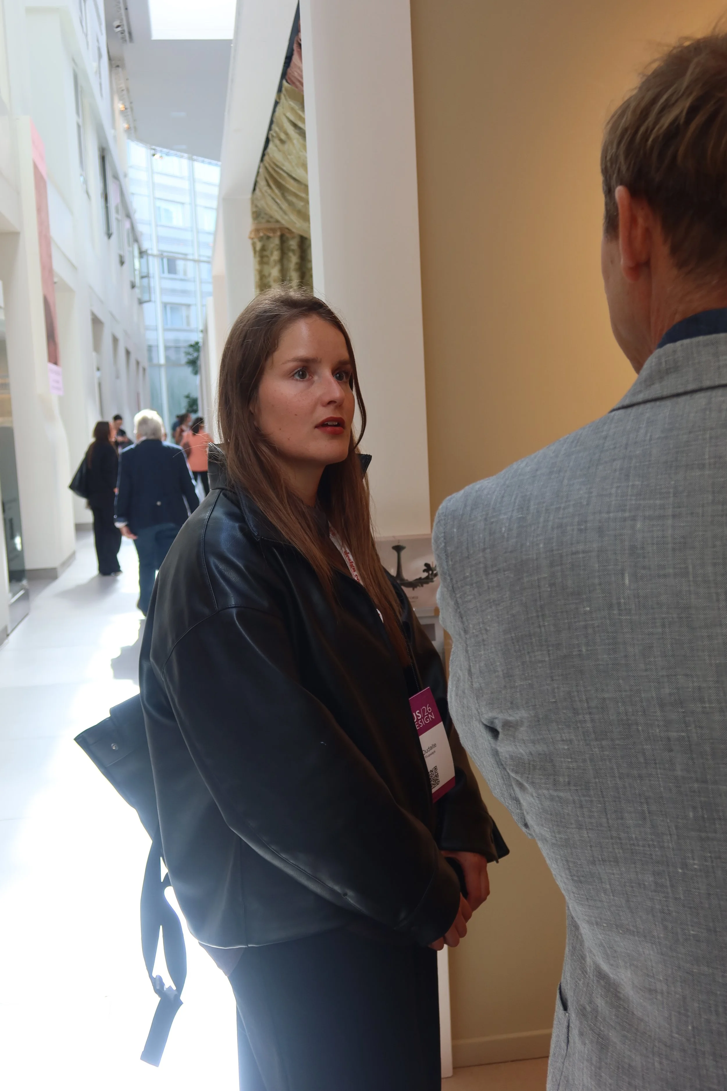 A young woman with long brown hair and a black leather jacket is engaged in conversation with a man in a gray suit inside a well-lit building. The woman looks attentive with her lips slightly parted. Several people are seen walking and conversing in the background.